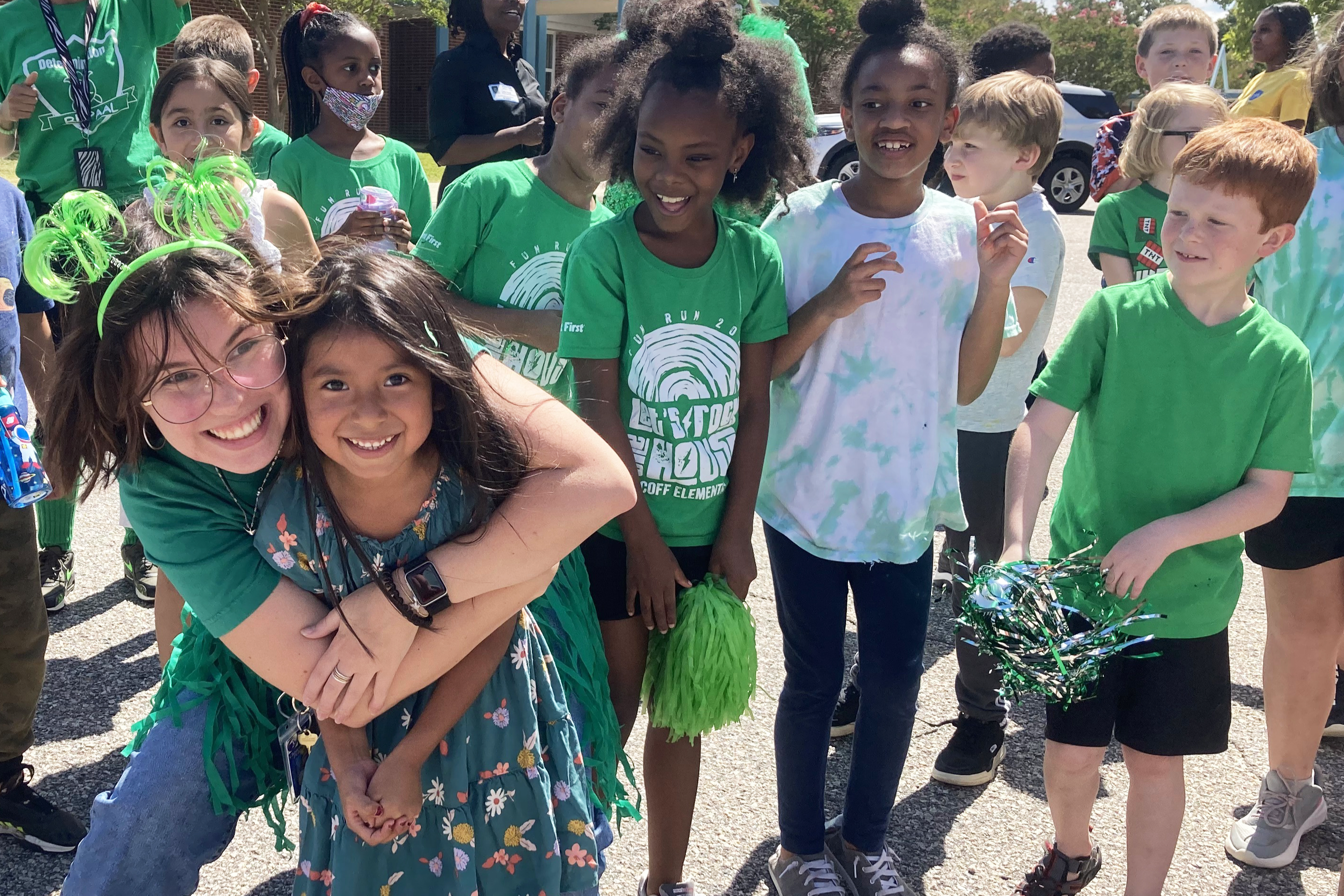 Students outside, most dressed in green. Teacher hugging one of them.
