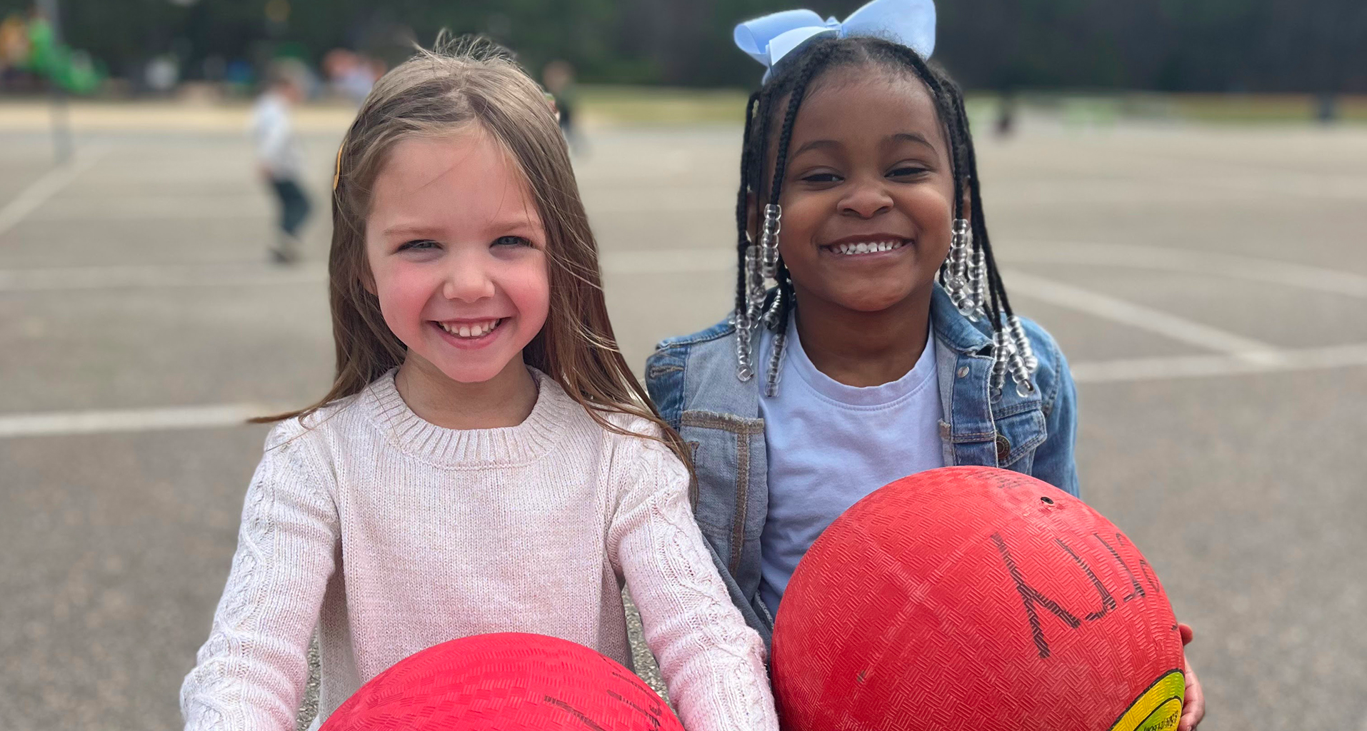 Two females on the playground holding big red balls smile for the camera.