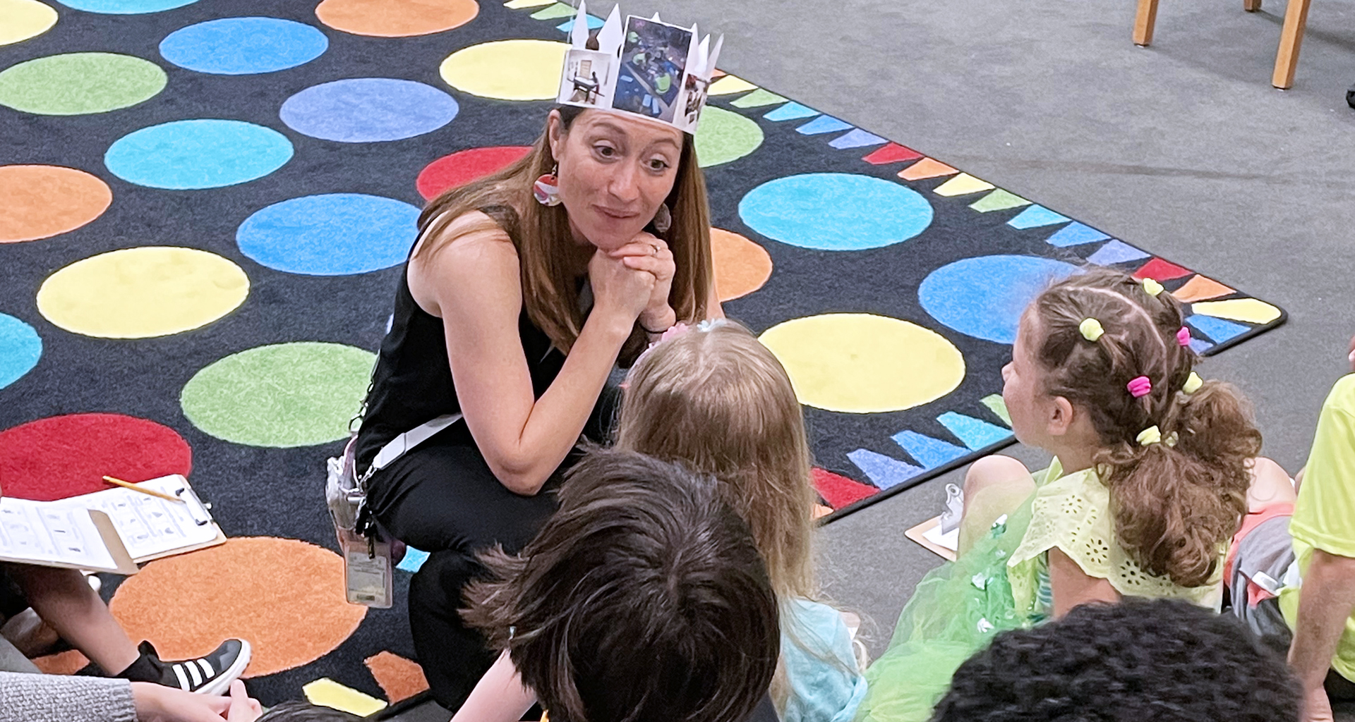 Teacher crouched down talking to students while wearing a paper crown.