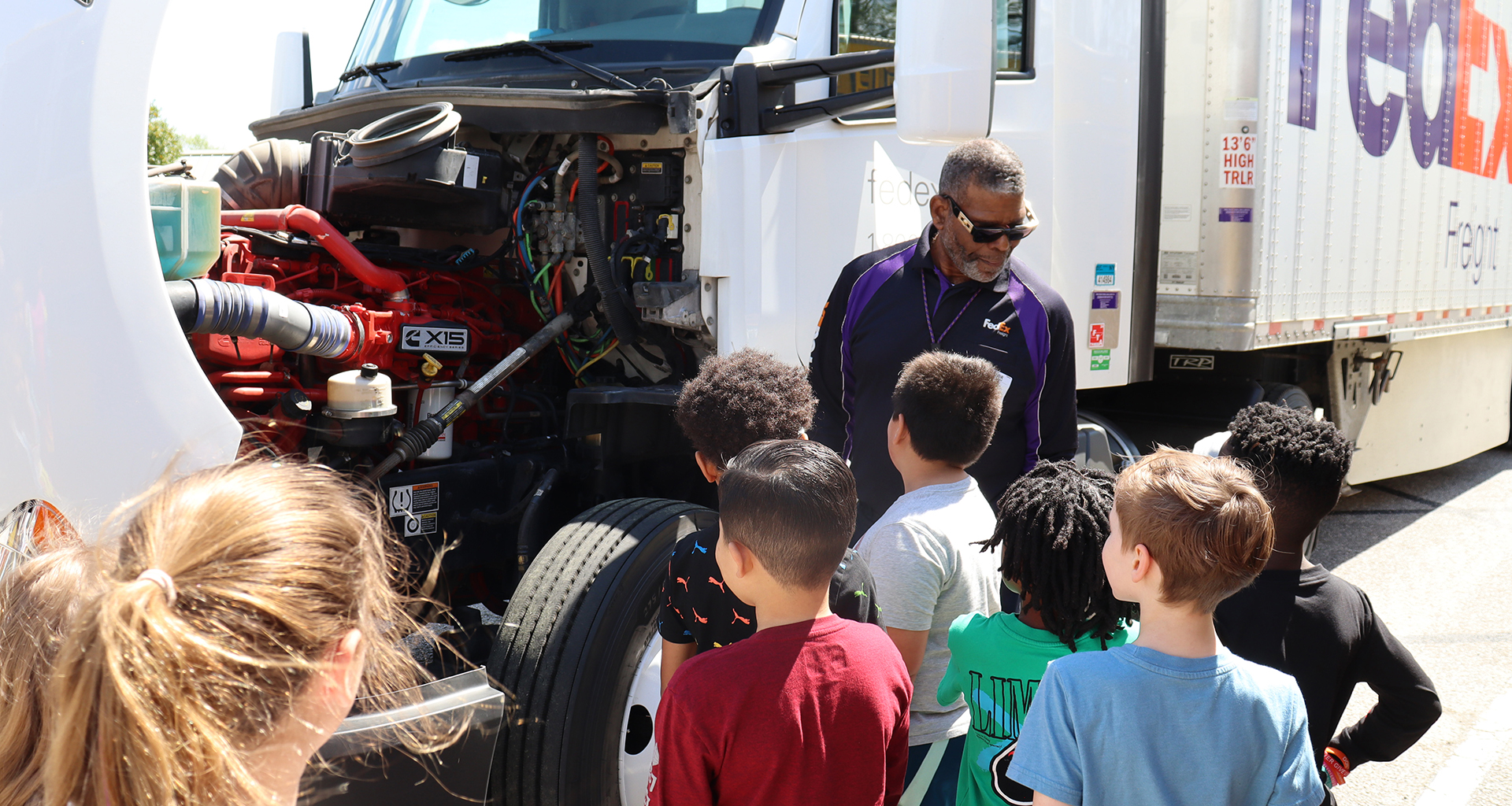 Several students looking at inside of a fedex truck engine while driver talks to them.