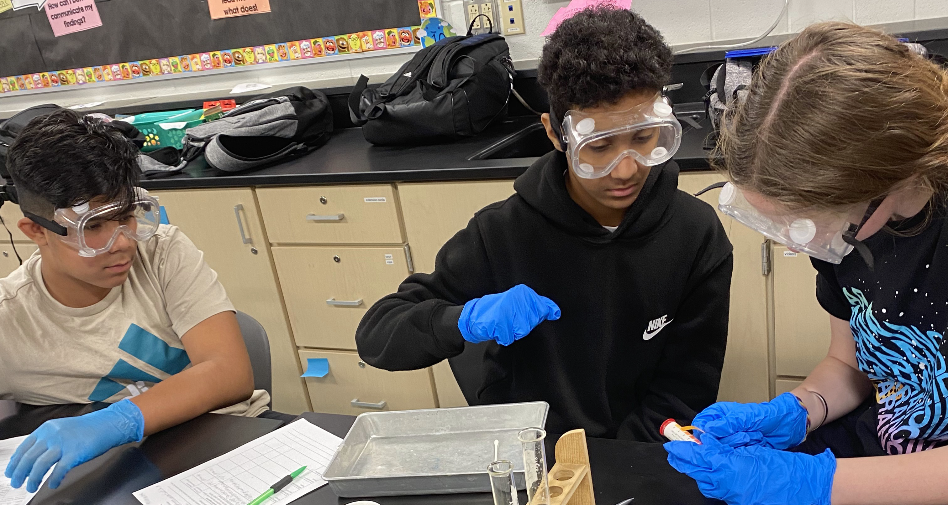 Three students wearing safety goggles and gloves in science class