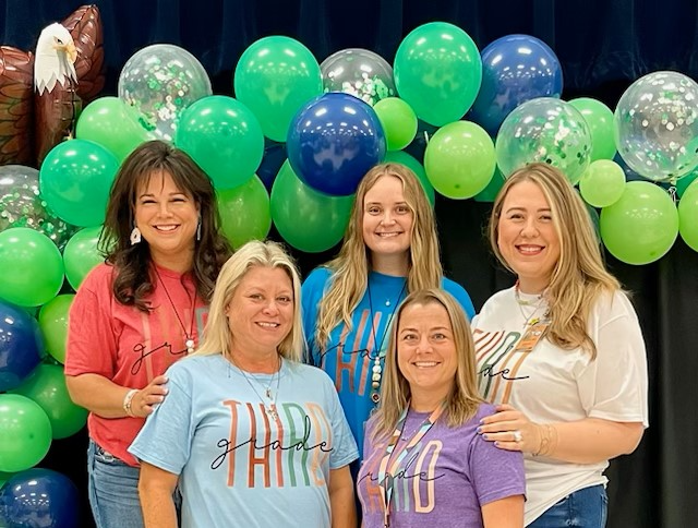 Third Grade teachers in gray shirts posing in front of blue wall 