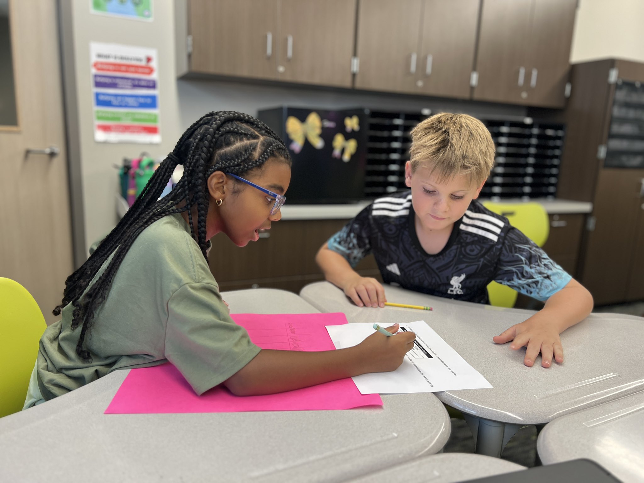 Two children seated at desks, one girl writing on paper, the other boy leaning over and pointing.