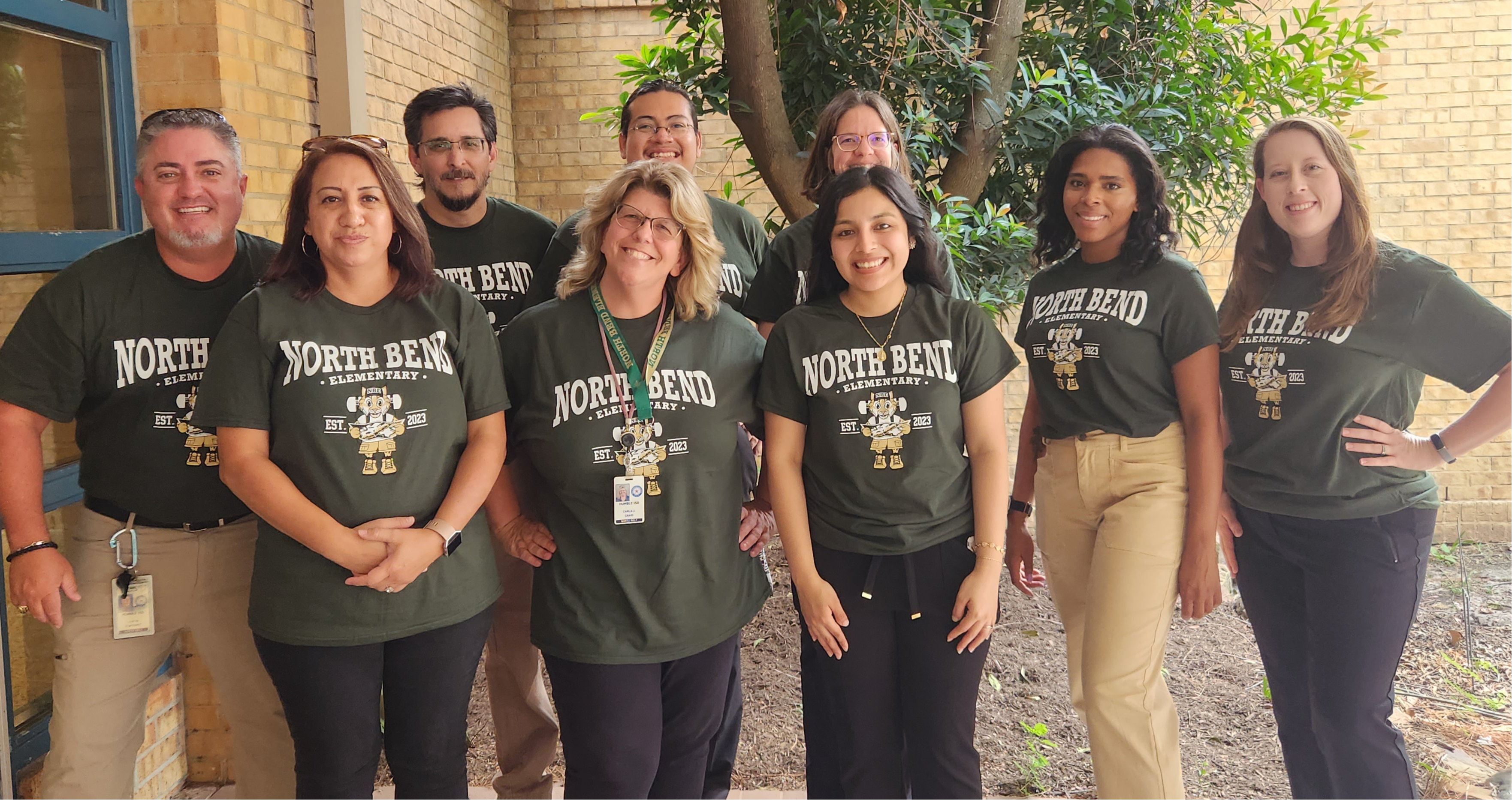 group of teachers in green shirts posing in front of tree outside school building