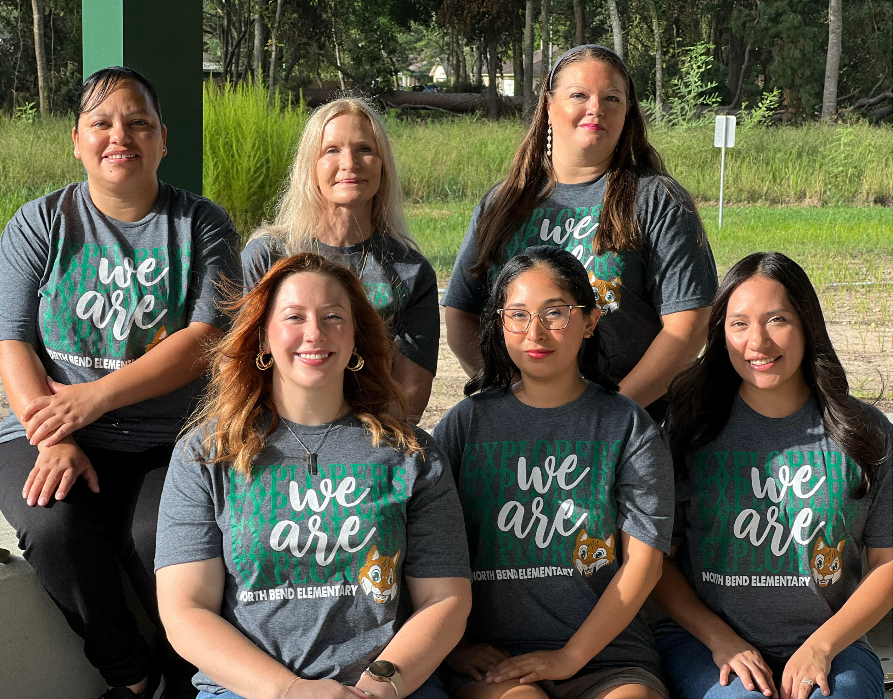 group of teachers in green shirts posing outside in front of greenery and building