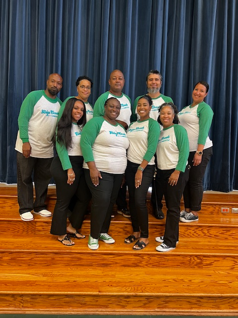 Fourth Grade teachers in blue shirts standing on stairs posing in front of stage curtain