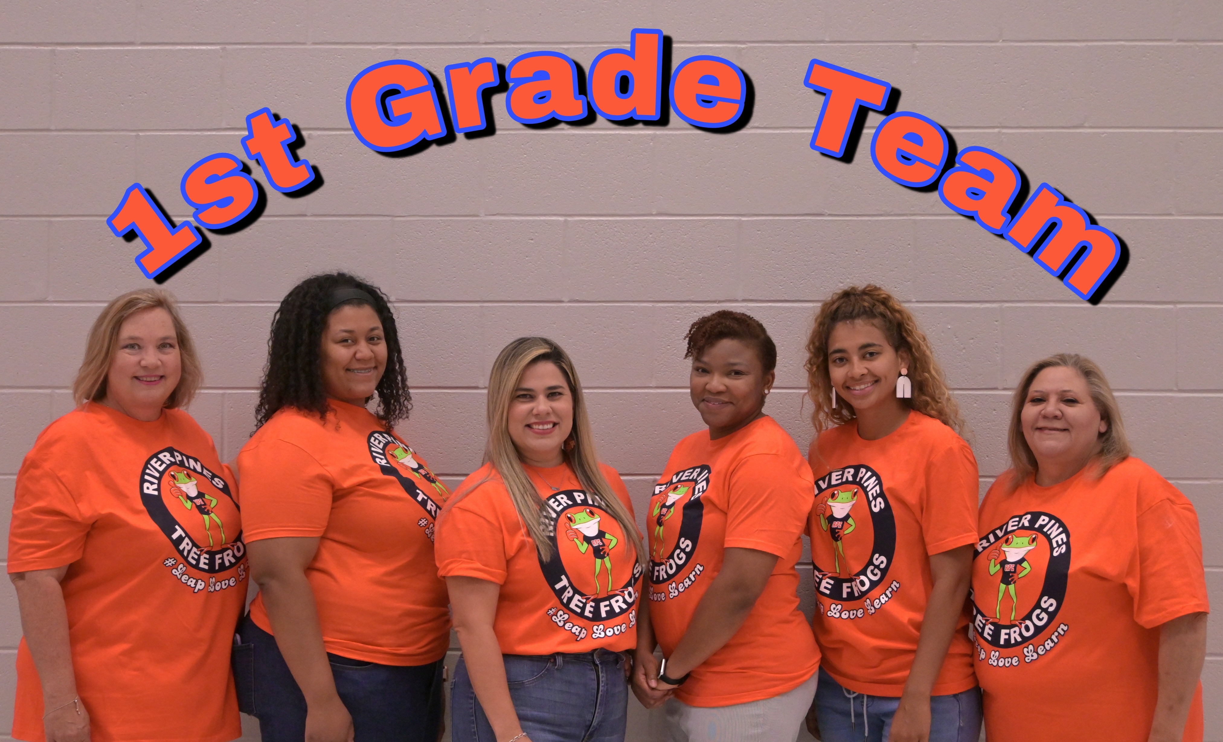 RPE 1st Grade teachers in orange shirts standing posing in front of gray brick wall