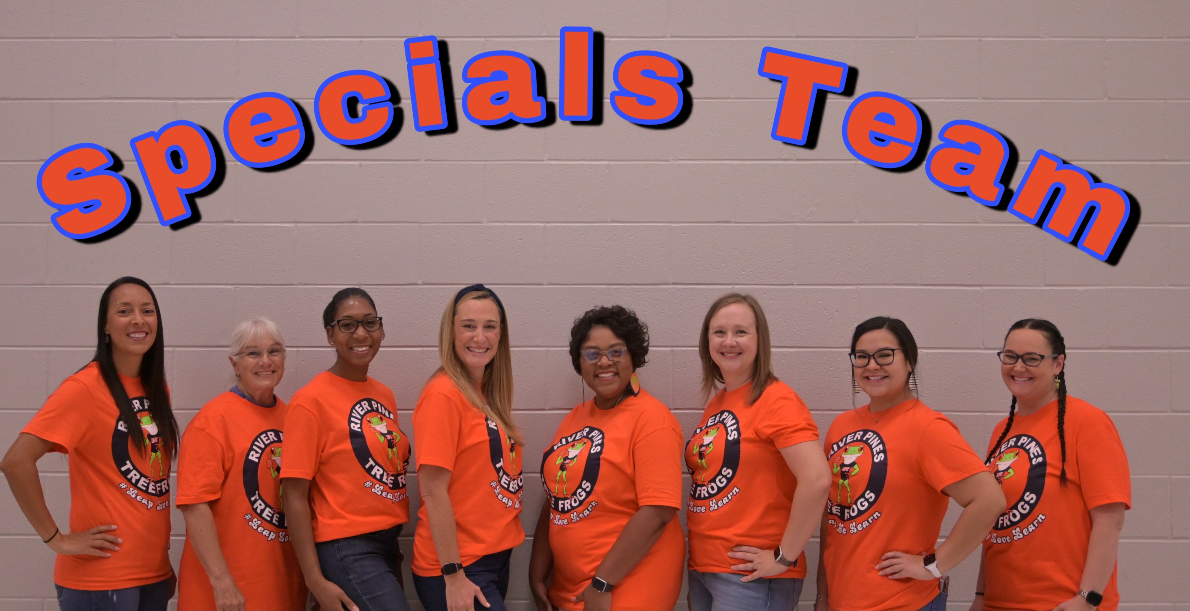 group of teachers in orange shirts standing posing in front of gray brick wall