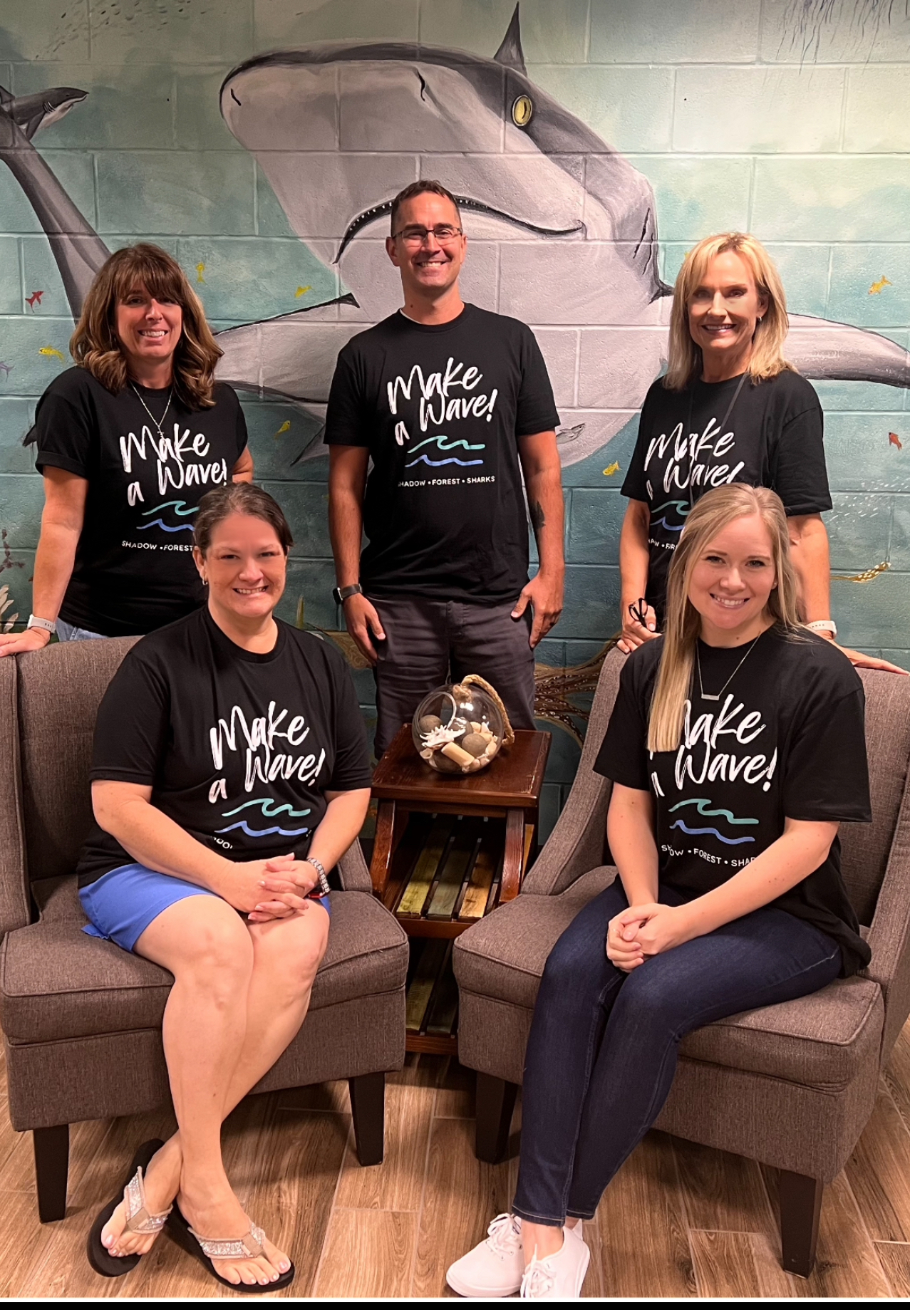 Fifth Grade teachers in blue shirts posing outside in front of window and brick wall