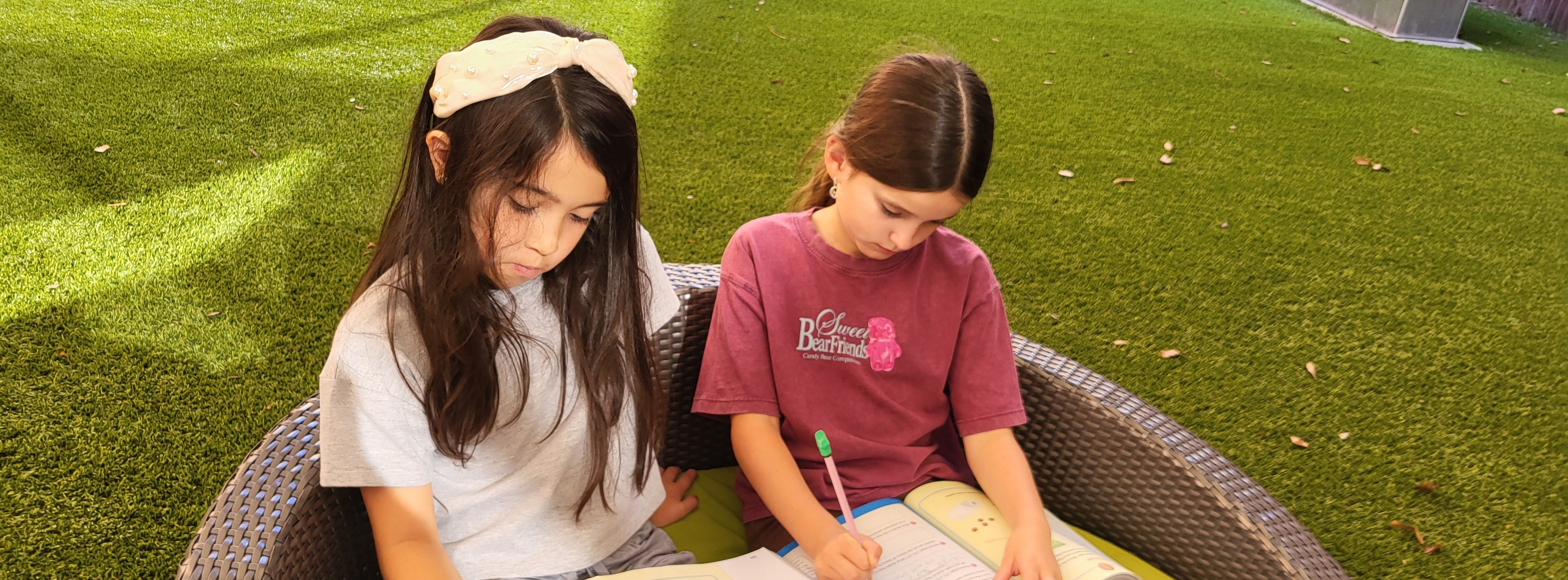 Two children seated on a round beanbag chair with books, pens, and colored pencils in a green grass area.
