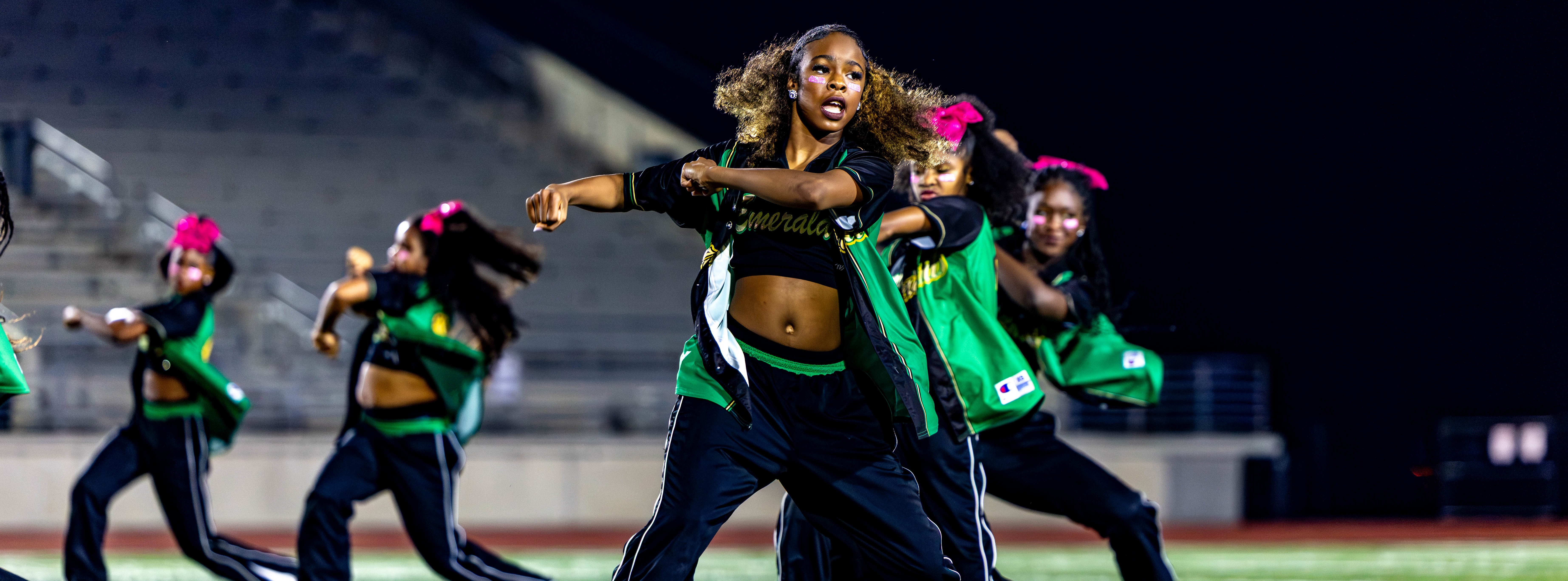 Group of dancers in green and black uniforms performing on a football field with bleachers in the background.