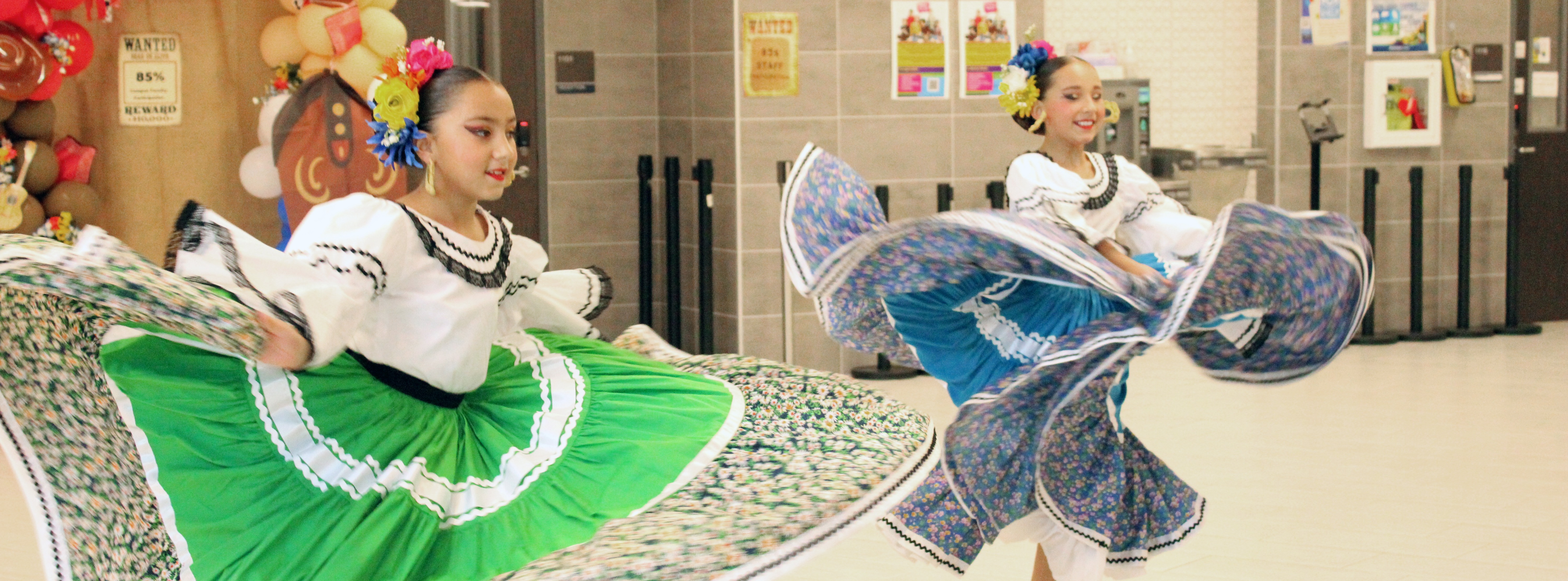 Two young girls in colorful dresses dance in a room with a tiled floor and decorations.