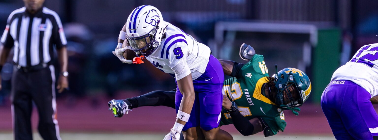Football players in action on a field, one in white and purple and one in green, with a referee standing.