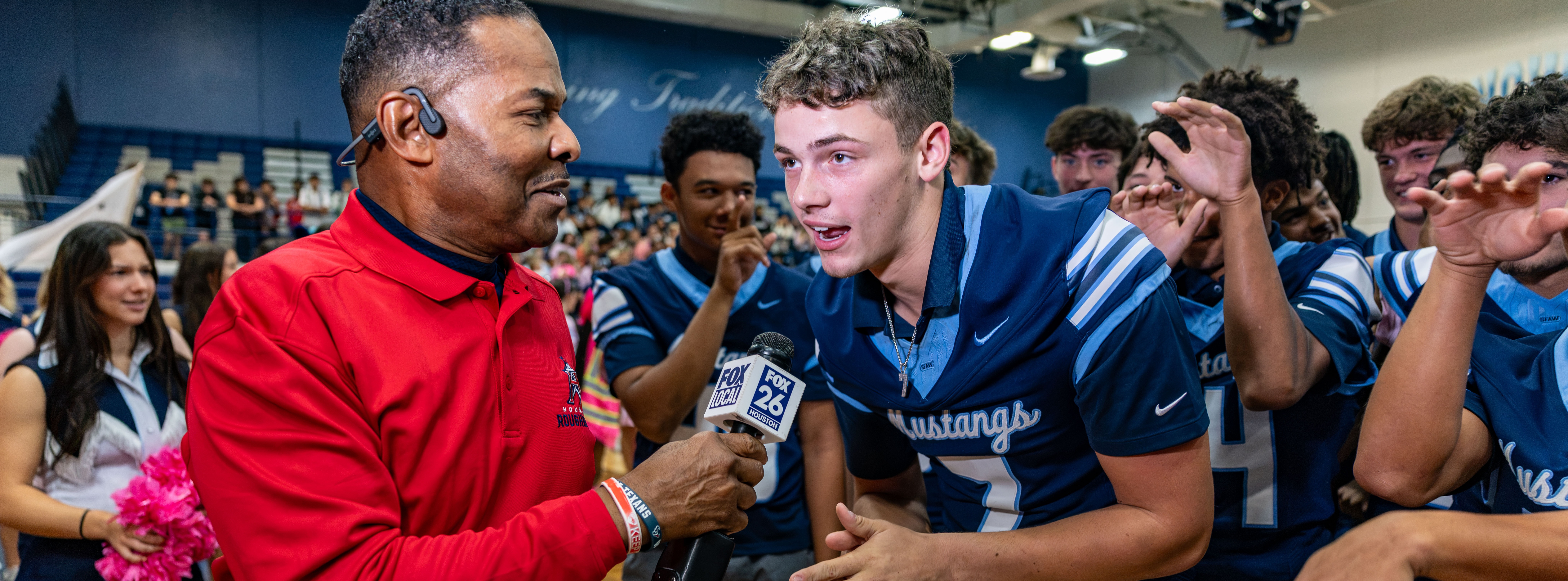 Man in red jacket interviewing boy in blue jersey on a basketball court with other students.
