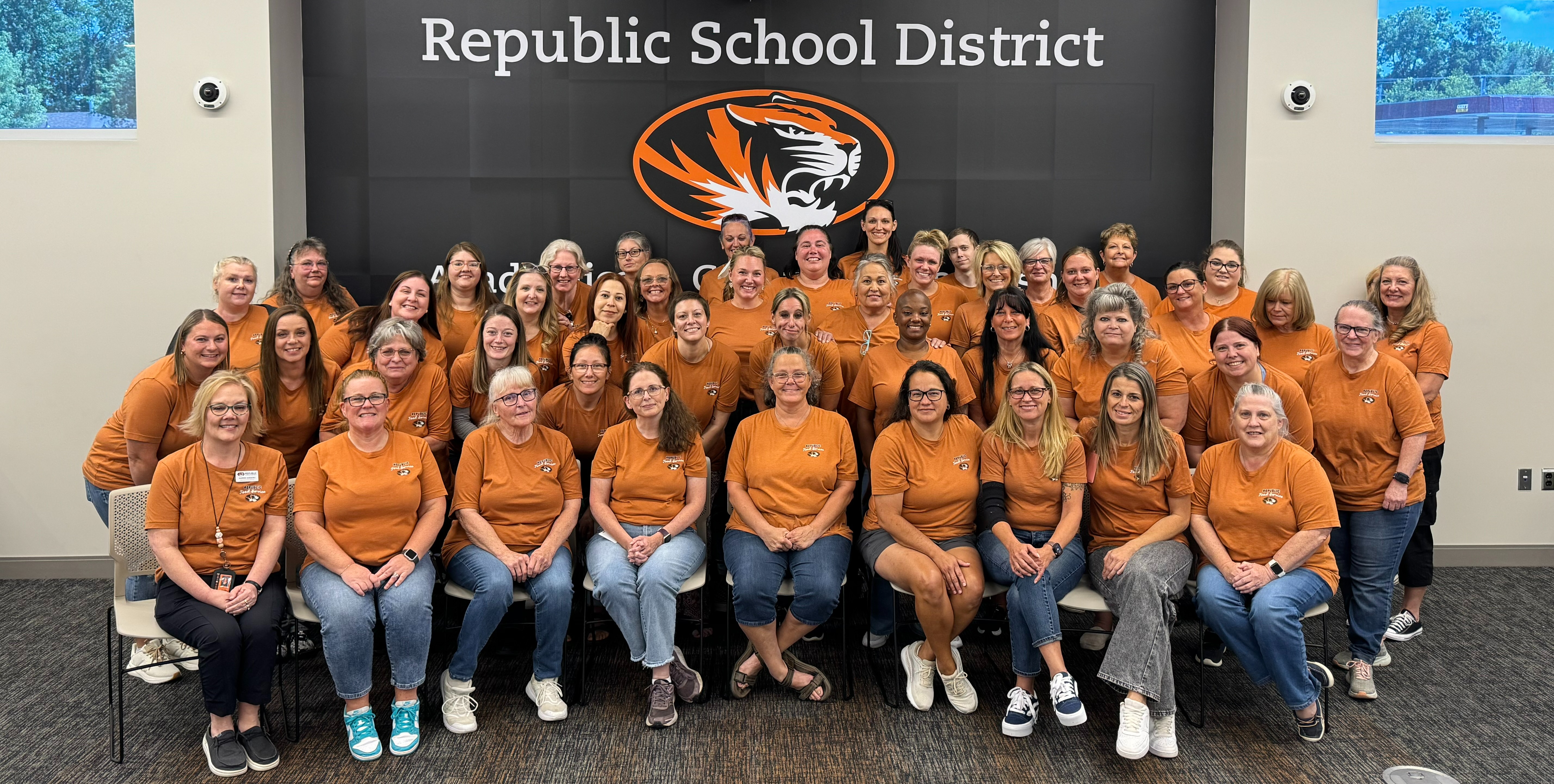 Members of the Food Service department standing and sitting together in a group smiling at the camera in front of a wall that says "Republic School District"