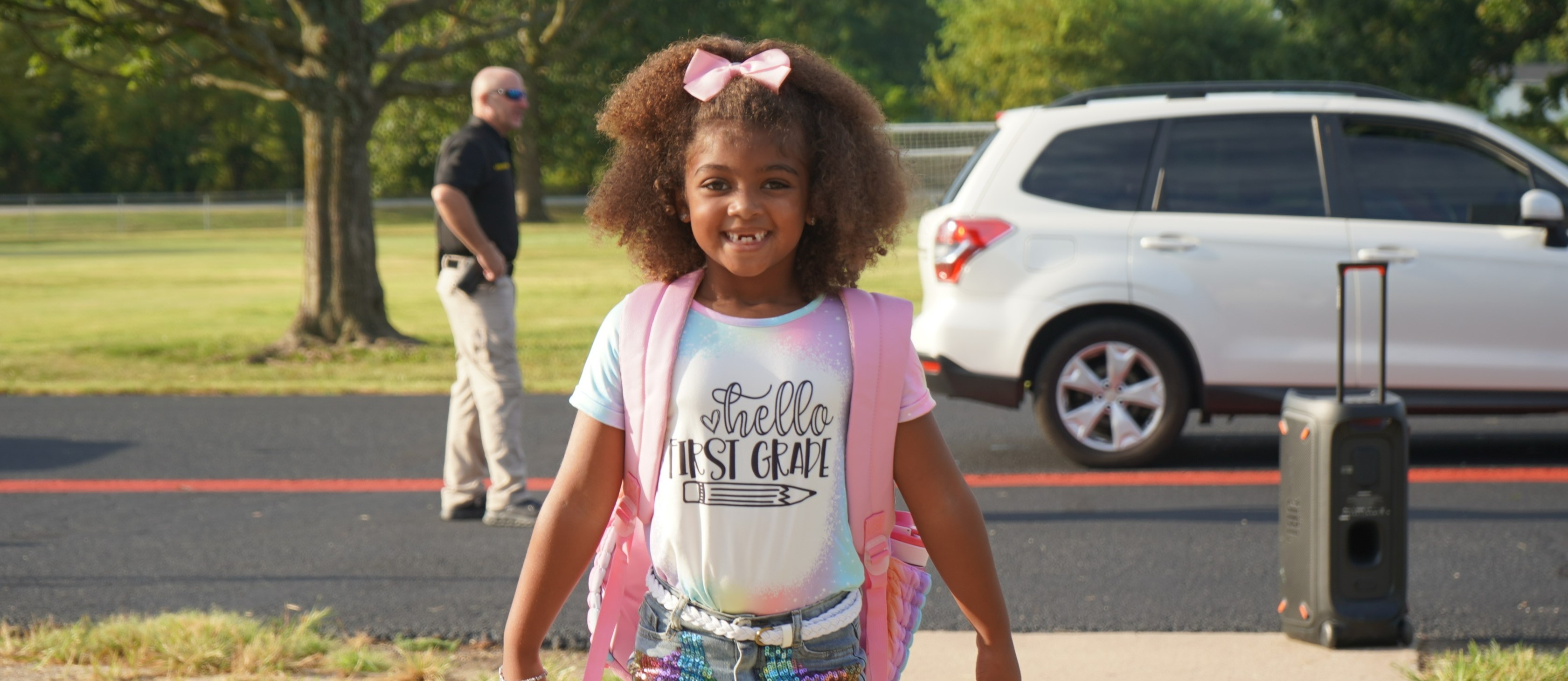 Student smiling, wearing a pink and blue shirt, wearing a pink backpack, walking into her school building