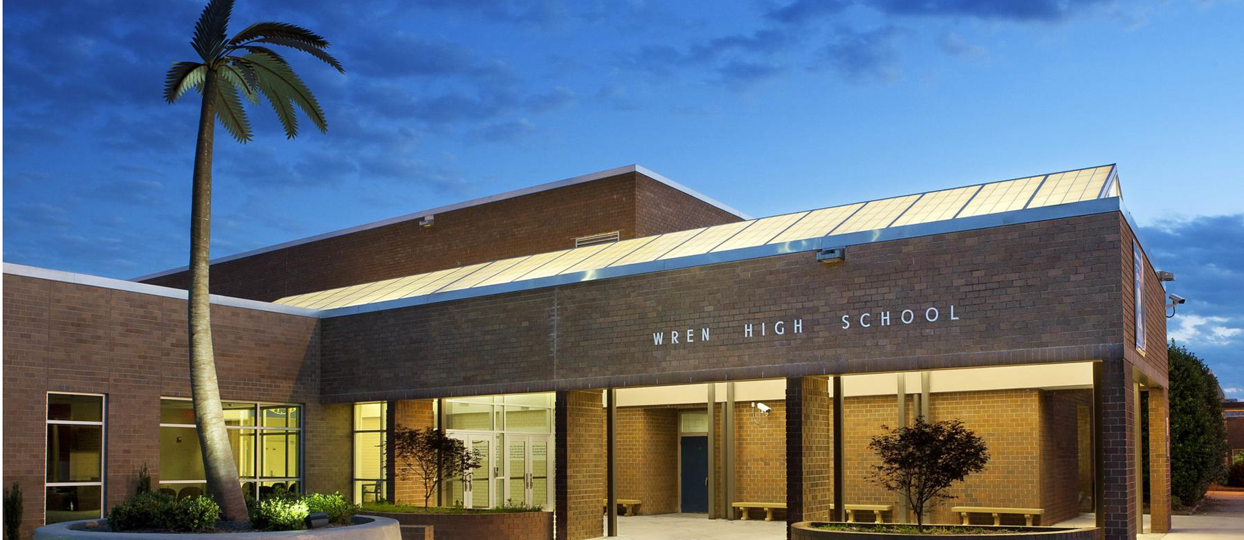 Photo of the front entrance of Wren High School.  A palm tree sets in front of a one story brick building that is lit up against a sunset sky.  The building letters read Wren High School.