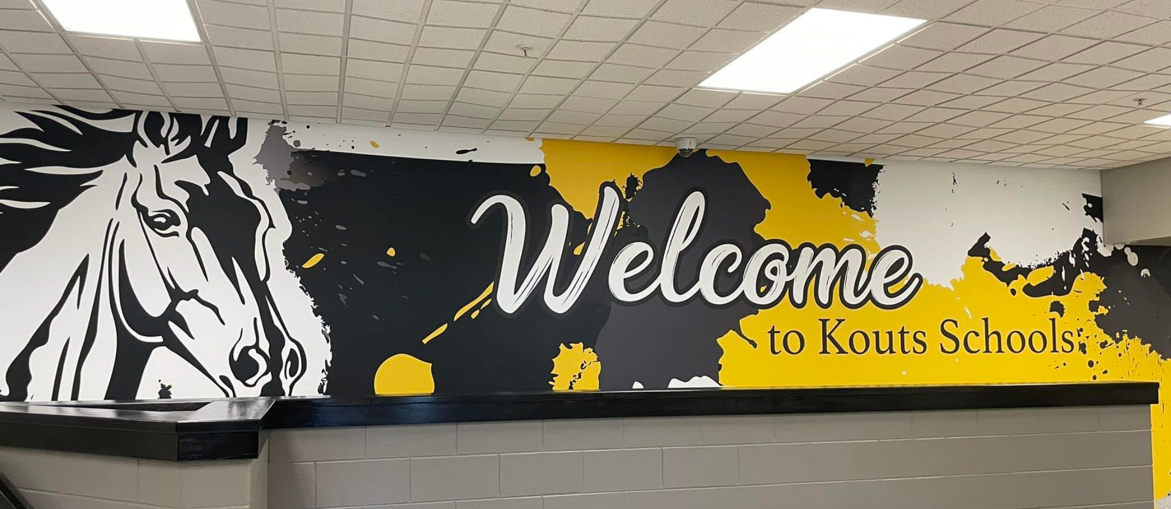 wall mural painted in black, gold, and white paint featuring head of mustang and stating "Welcome to Kouts Schools"