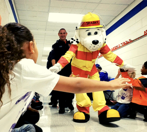 Mascot of a dog dressed like a firefighter gives children high-fives in hallway
