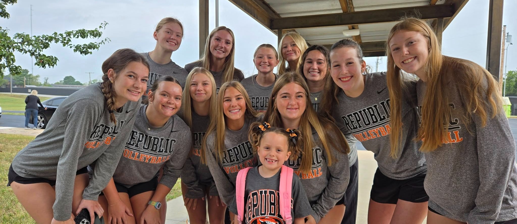 Student with pigtails and pink backpack  standing in front of 12 high school volleyball players in gray shirts and black shorts.
