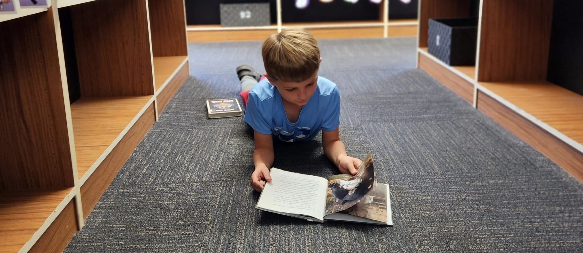 Student with light hair and blue t-shirt reading a book in the floor of the library.