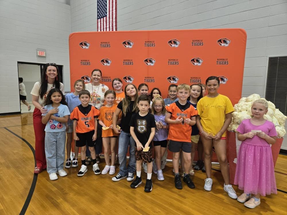Two female teachers with several students in front of an orange backdrop smiling for the camera.