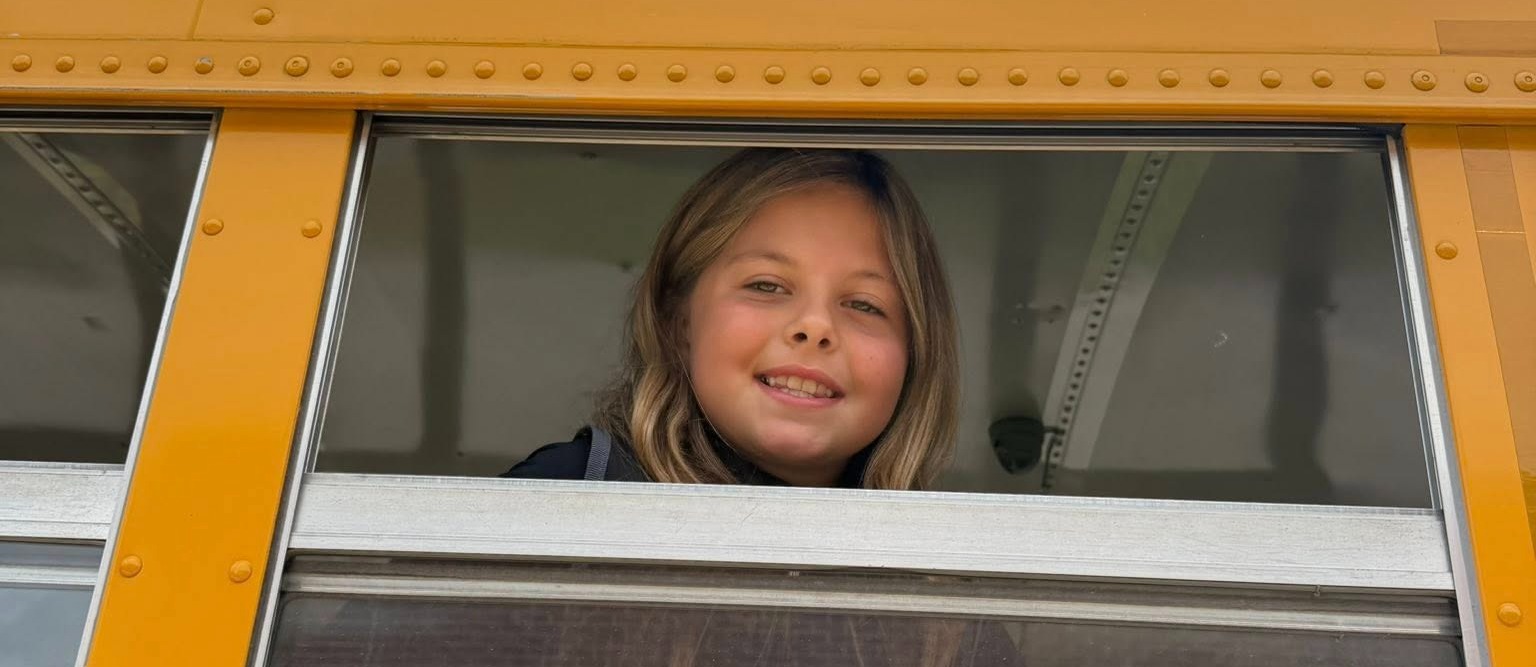 Student with light hair smiling out of the bus window.