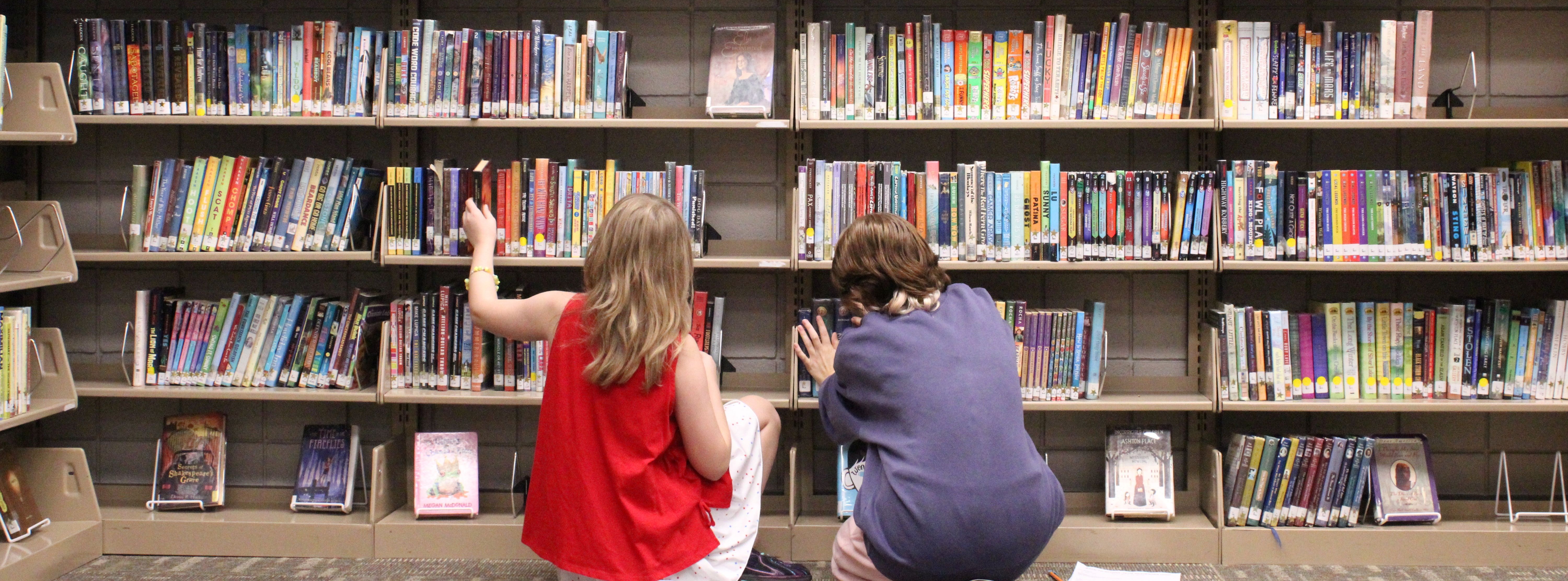 two students sitting on the ground infront of a large bookshelf of books
