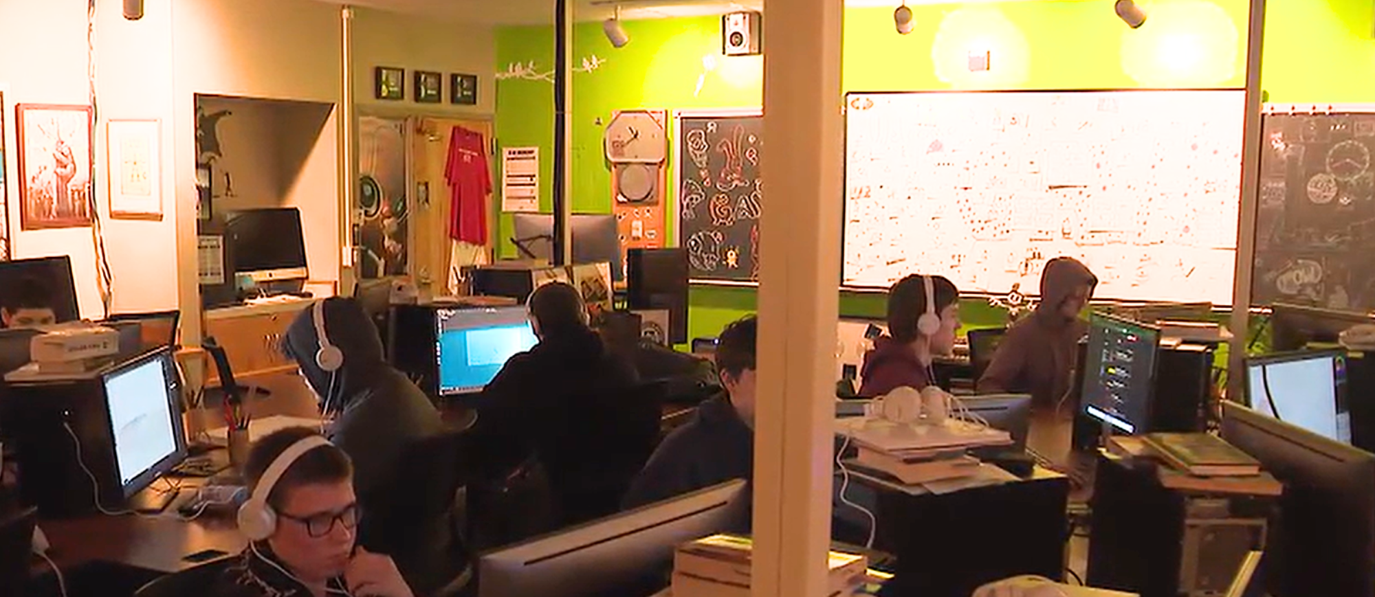 A room with green walls holds a group of people working on computers. The room has monitors, desks, and a whiteboard.