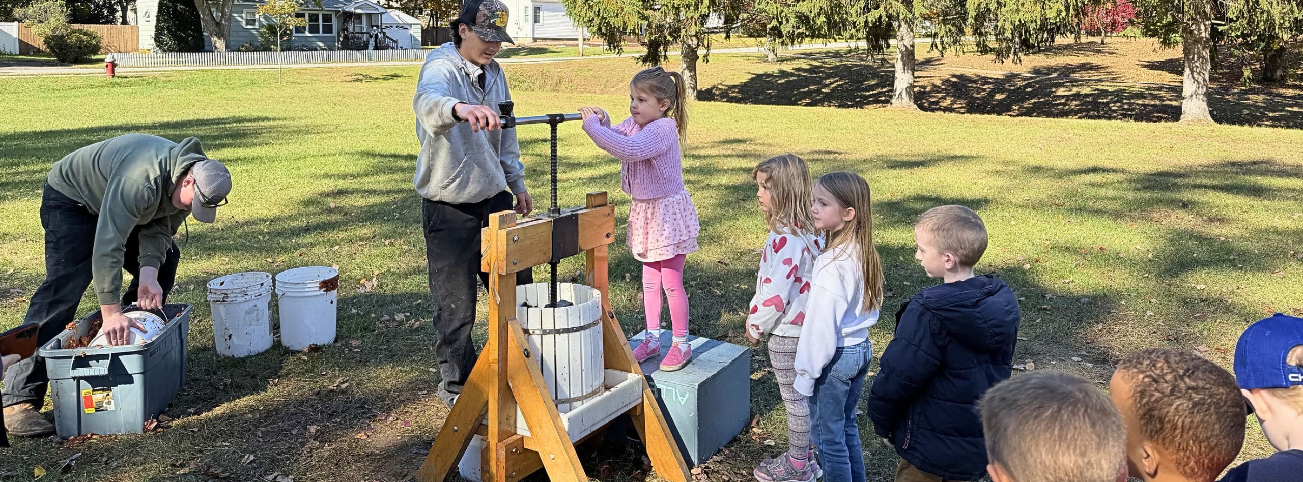 People gathered on a field, children using a wooden apple press, two adults operating it, buckets nearby.