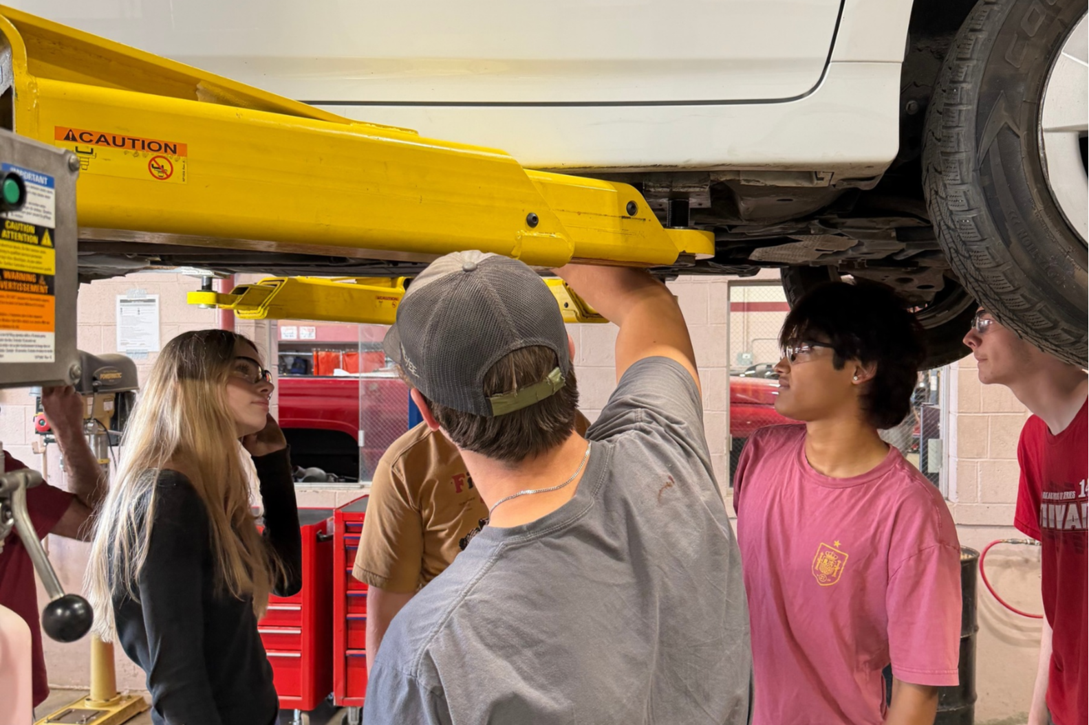 Students observe a car lifted by a yellow jack under a red brick wall.
