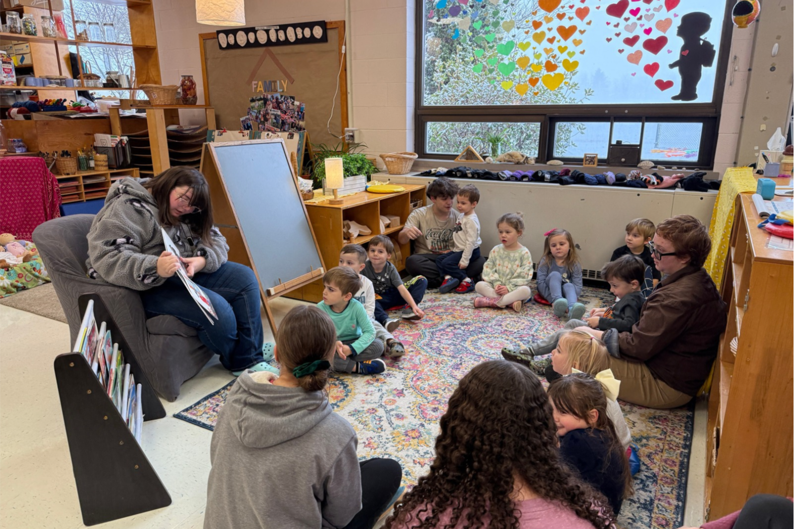 Classroom with children sitting in a circle on a rug. Two adults sit with them.