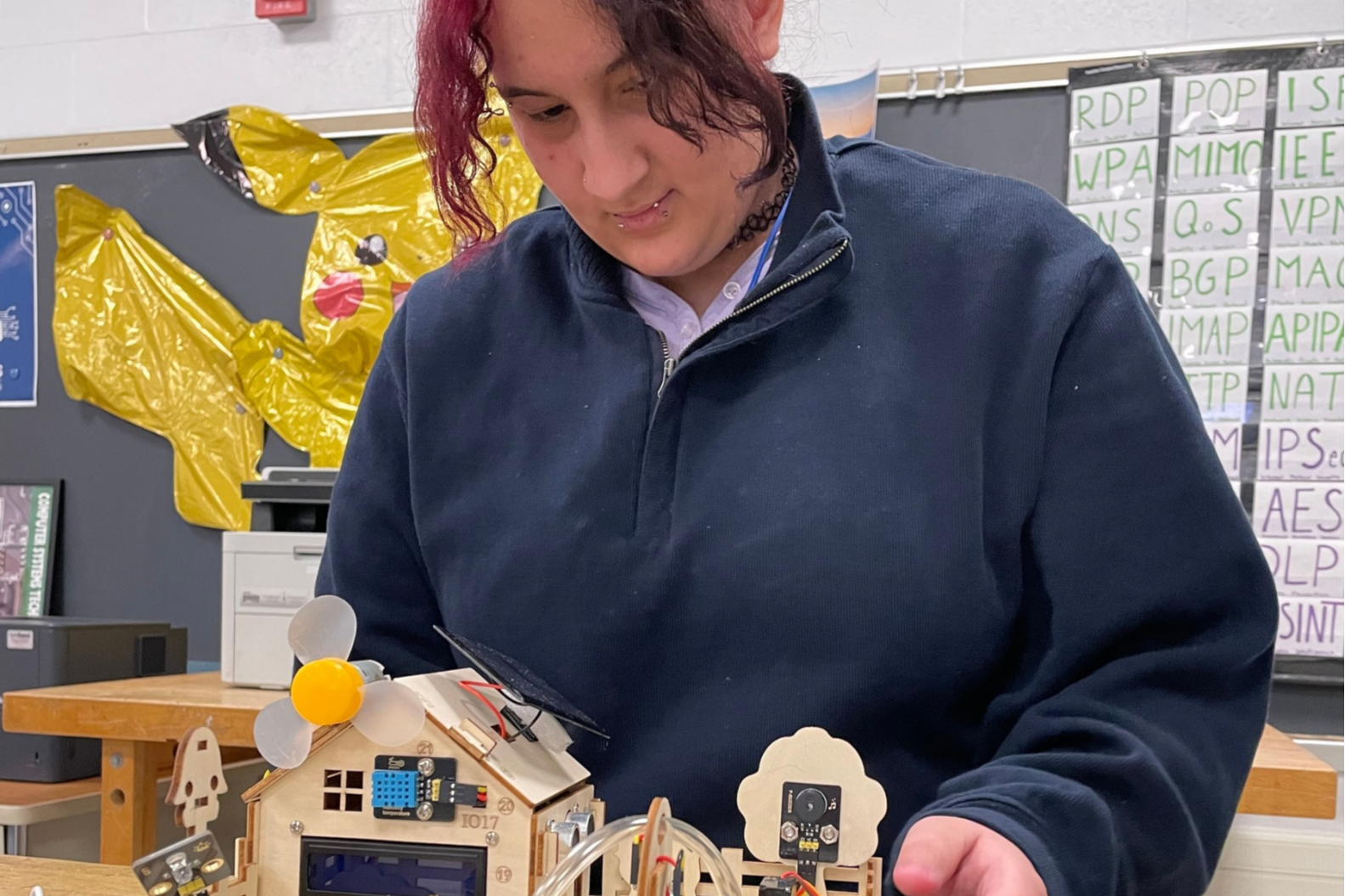 Person examining a miniature model of a house and solar panel. Yellow Pokemon balloon in the background.