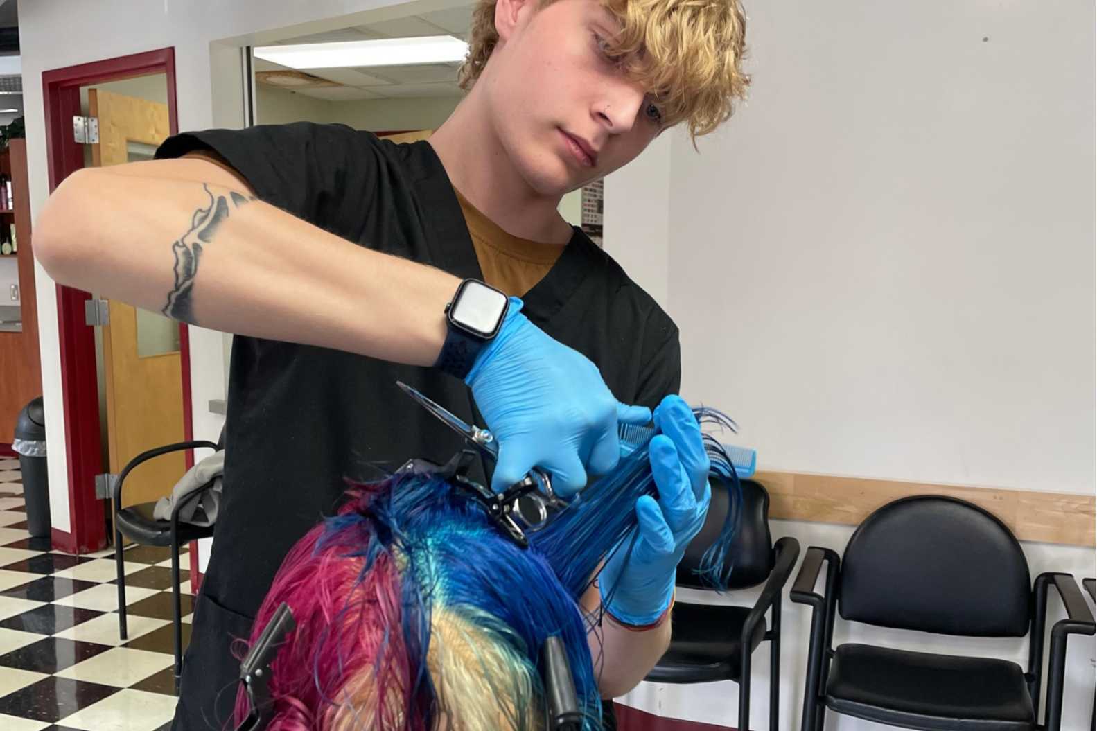 A person with tattoos and a watch cuts hair on a mannequin head with scissors.