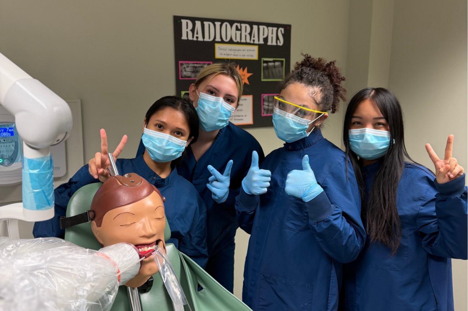 Five students in blue uniforms and masks stand behind a dental mannequin, making gestures with their hands.