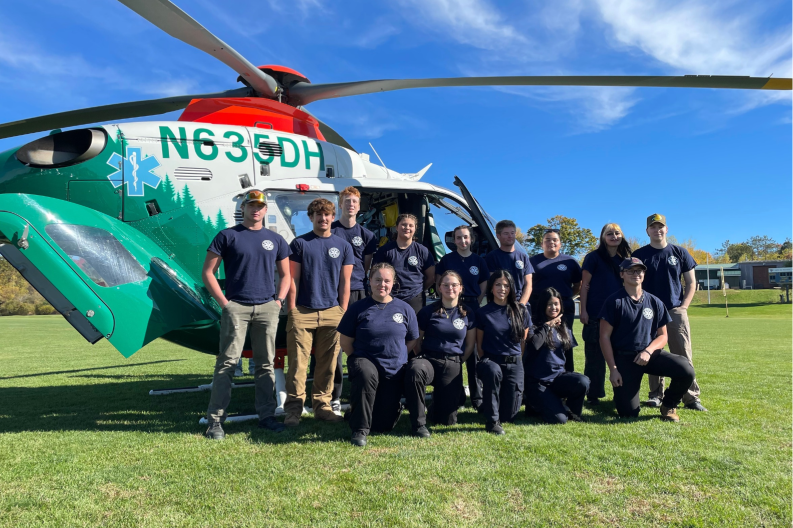 A group of people in blue uniforms, some sitting and some standing, pose in front of a green and white helicopter.