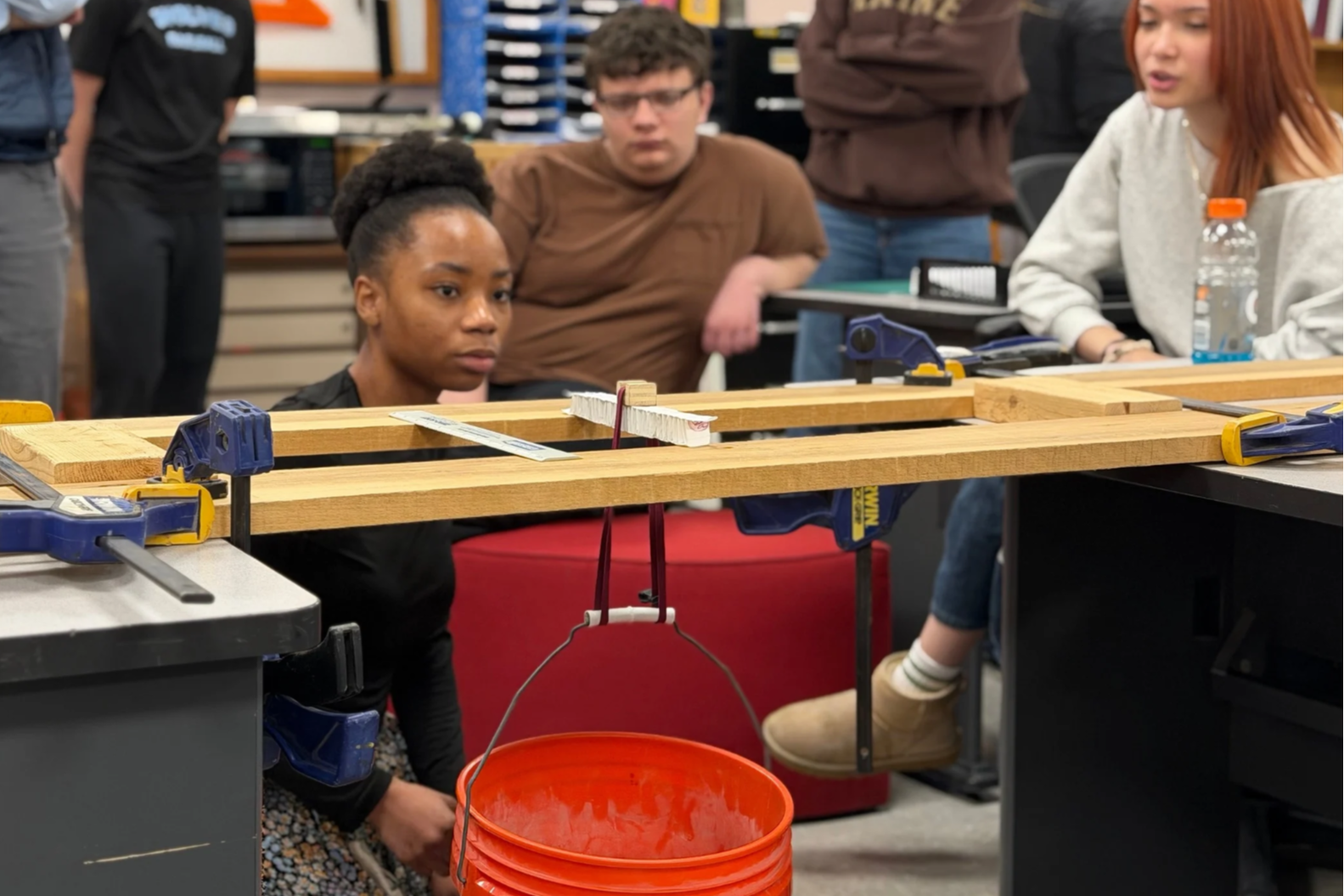 Students work on a wooden beam, one holding a bucket, others standing around. One student kneels in front of a counter.