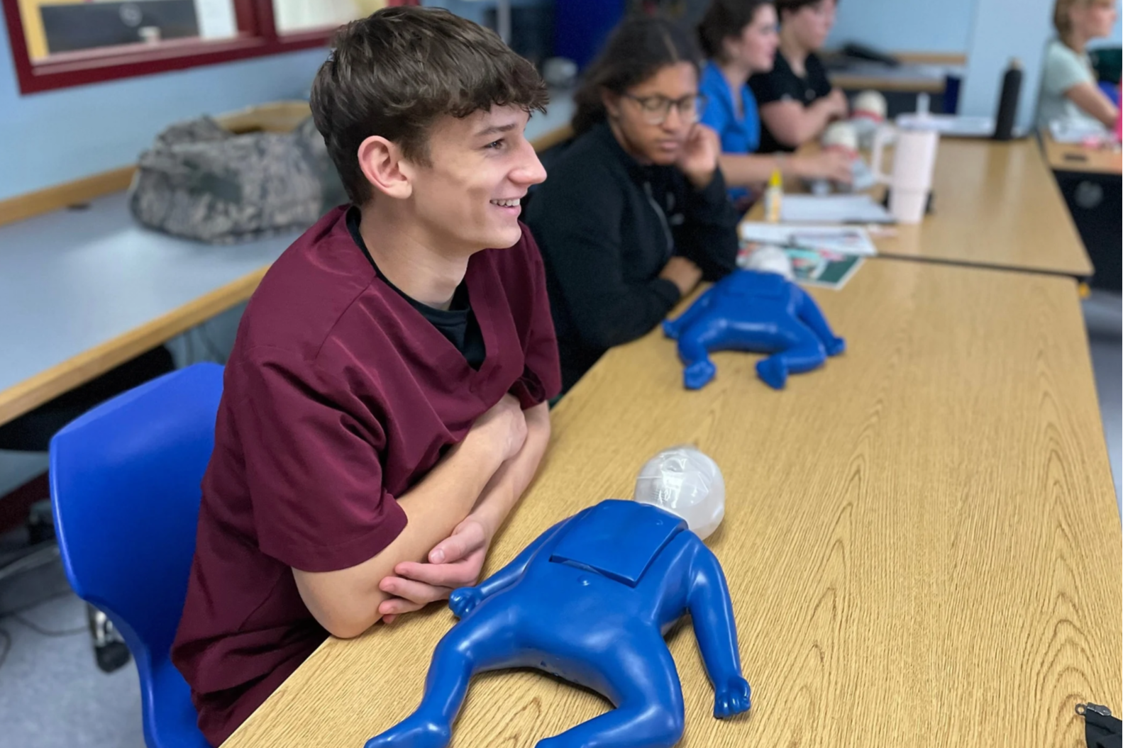 Several students sit in a classroom, one boy smiles. Blue mannequins are on the table.
