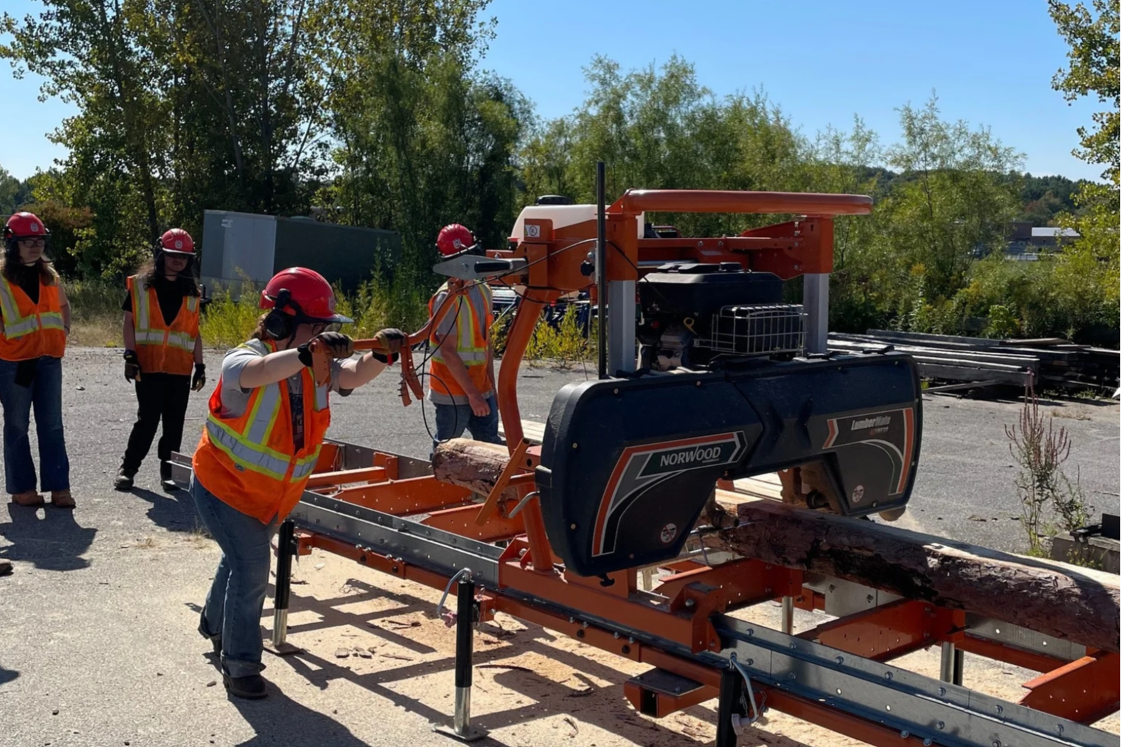 Five people in orange vests and helmets using a saw to cut wood outdoors on a sunny day.