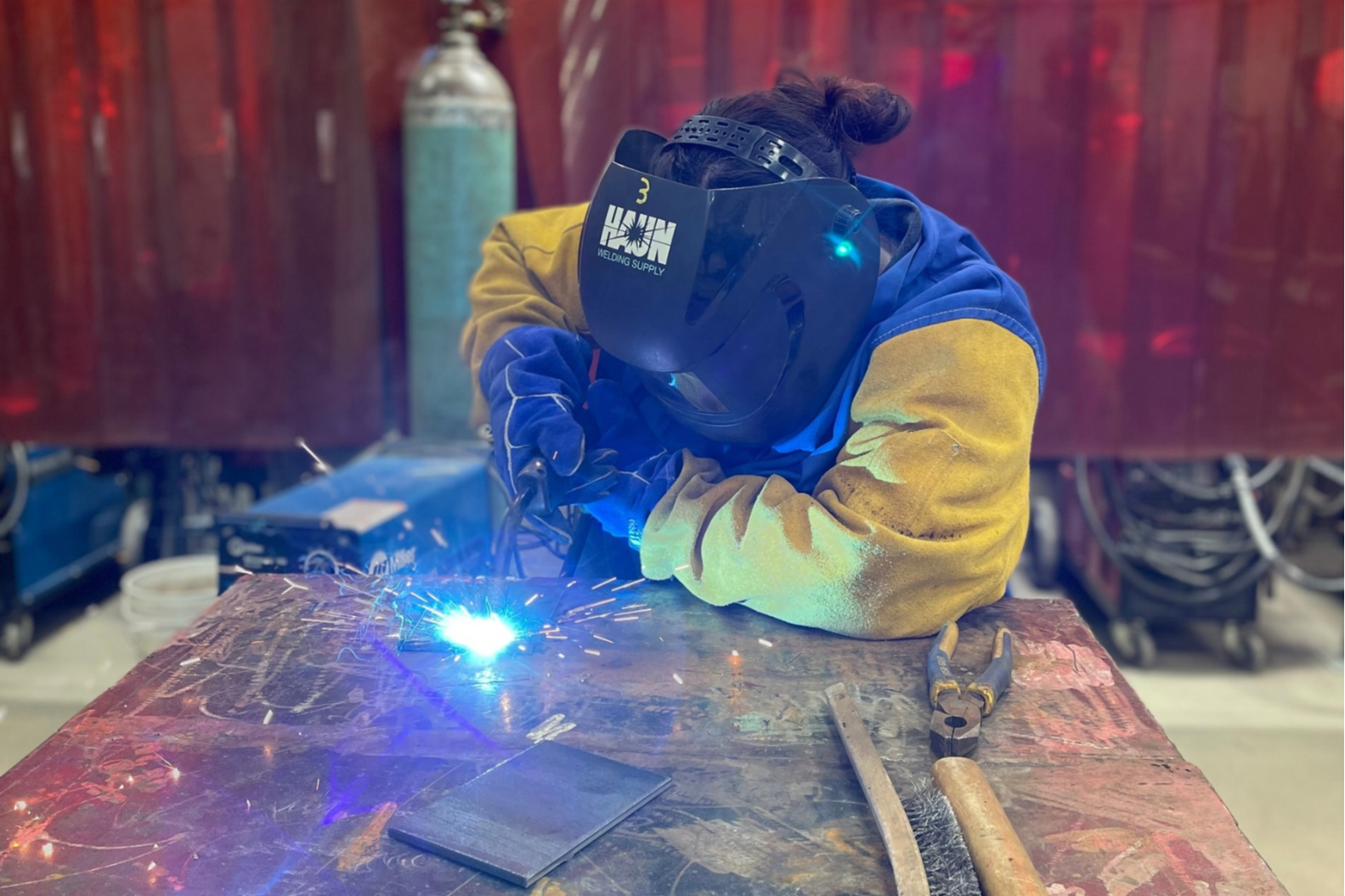 Person in welding gear welding metal on a table with tools and a red wall in the background.