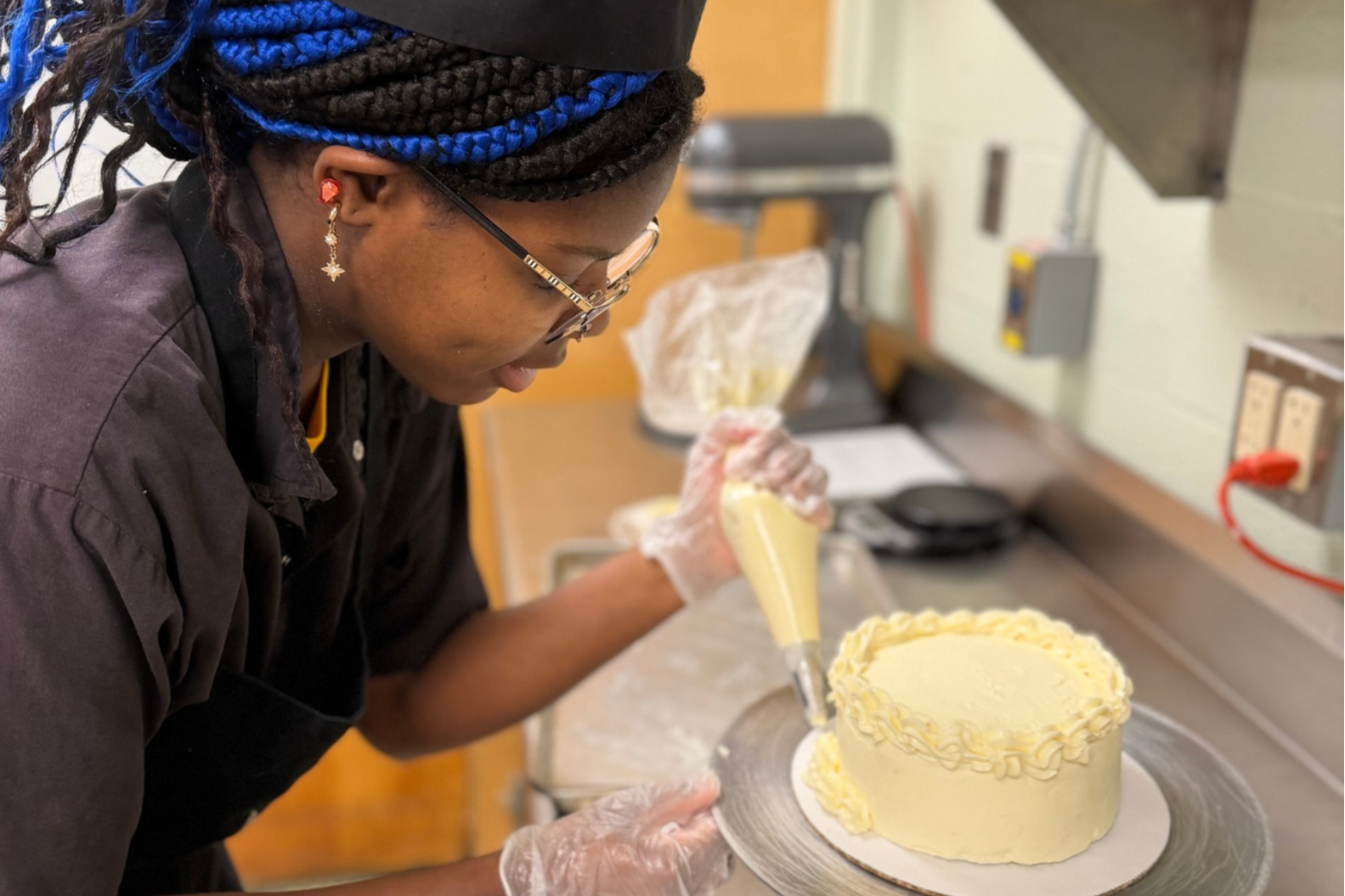 Person decorating a cake in a kitchen with a cake stand, piping bag, and cake.