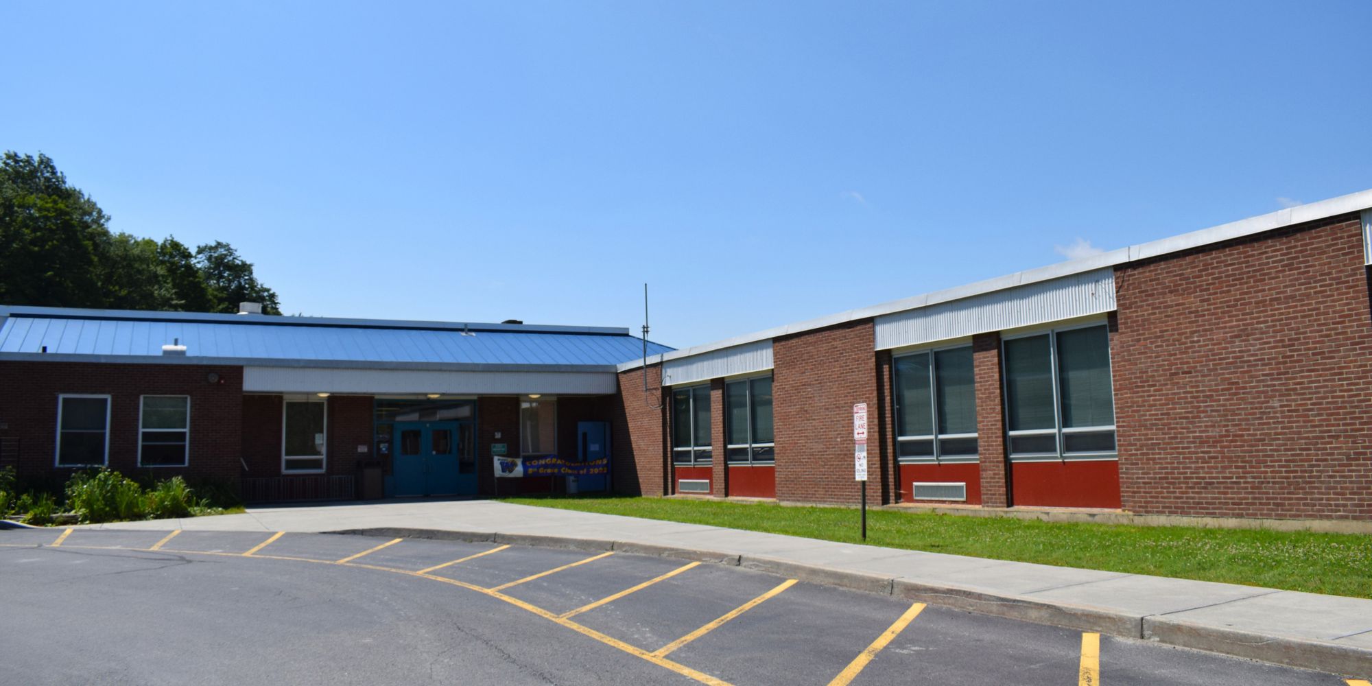 A school building with red and brown brick walls. Large windows, a blue roof, and a grassy area in front.