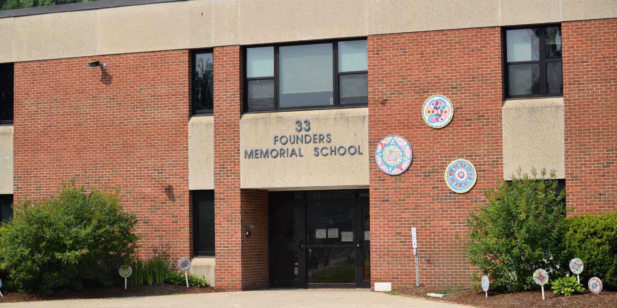 Brick school building with glass windows, "33 Founders Memorial School" in large letters above the entrance. Two round emblems on either side.