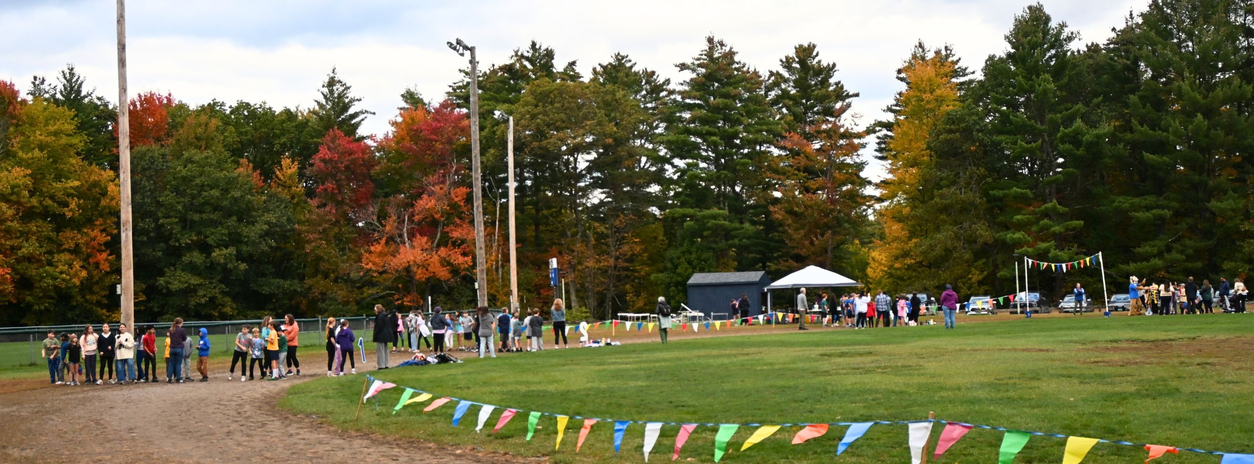 An open field with people gathered. Colorful banners line the sides. Trees with autumn leaves in the background.