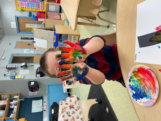 A child holds up his hands which are painted rainbow colors.