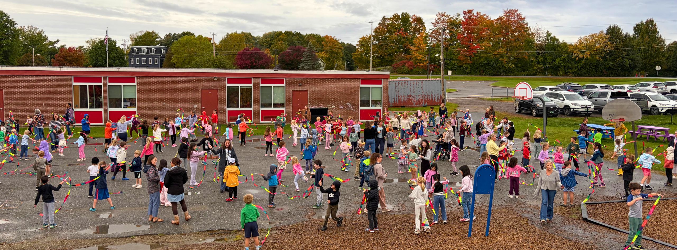 Crowd of children in bright colors standing in a field, with a red school building and cars in the background.