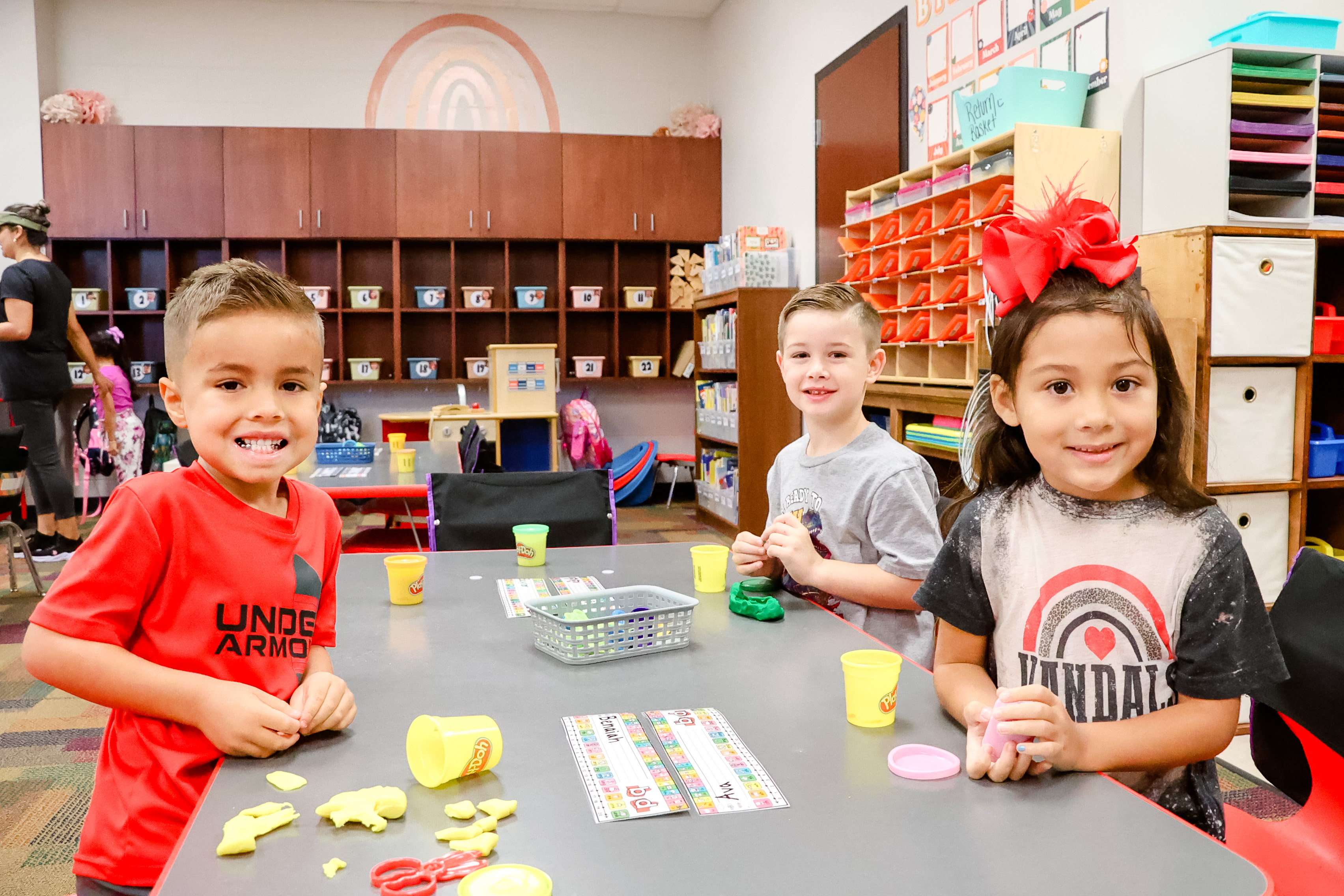 three students smiling at their desk playing with play doh