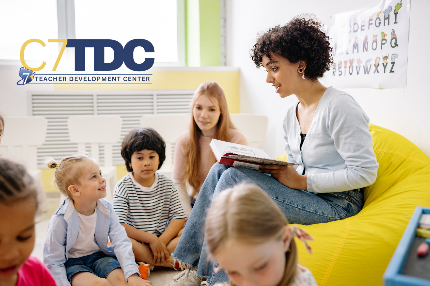 Teacher sitting in front of young students on the floor, reading from a book.