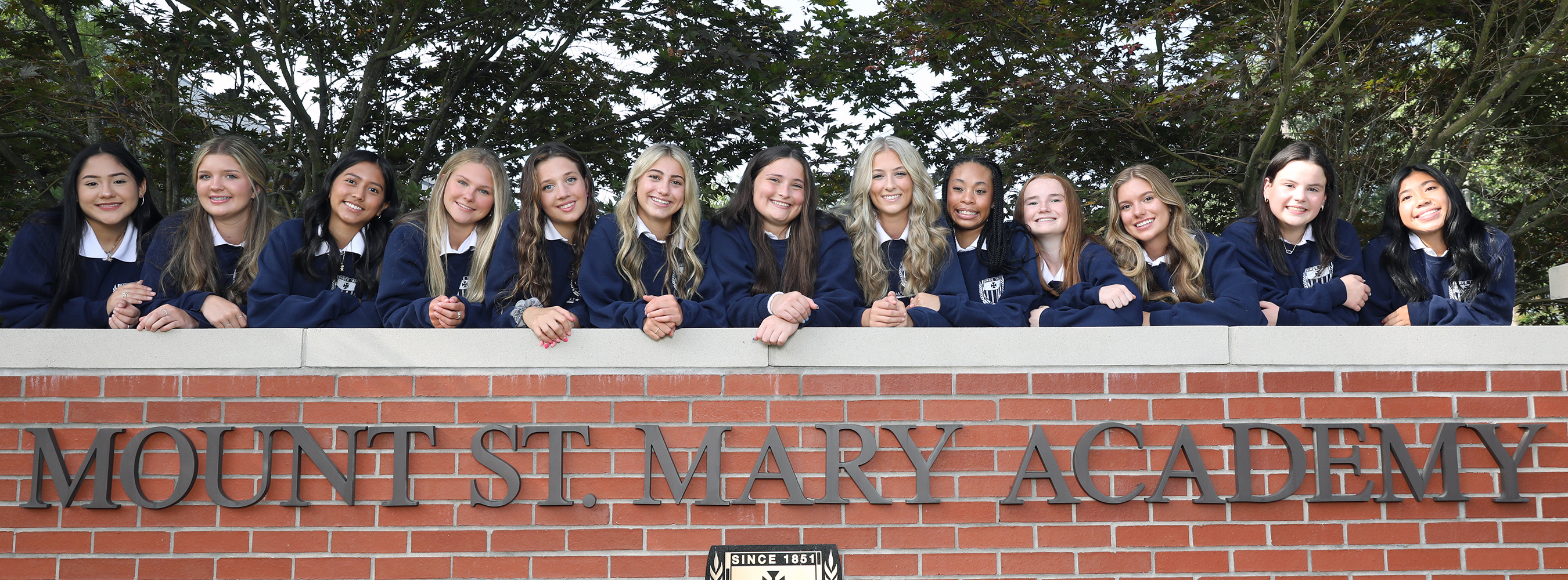 large group of students standing at the brick Mount St. Mary Academy sign and smiling