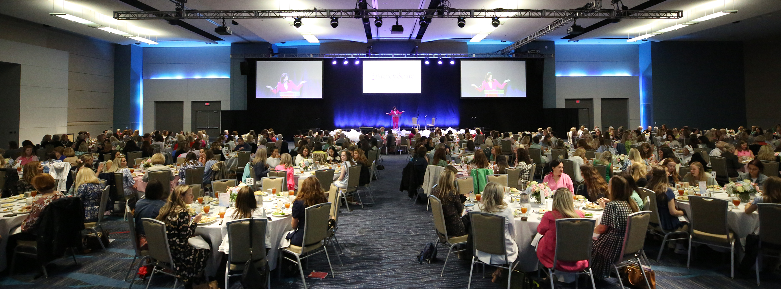 ballroom full of tables and seated guests listening to a speaker presenting from a stage